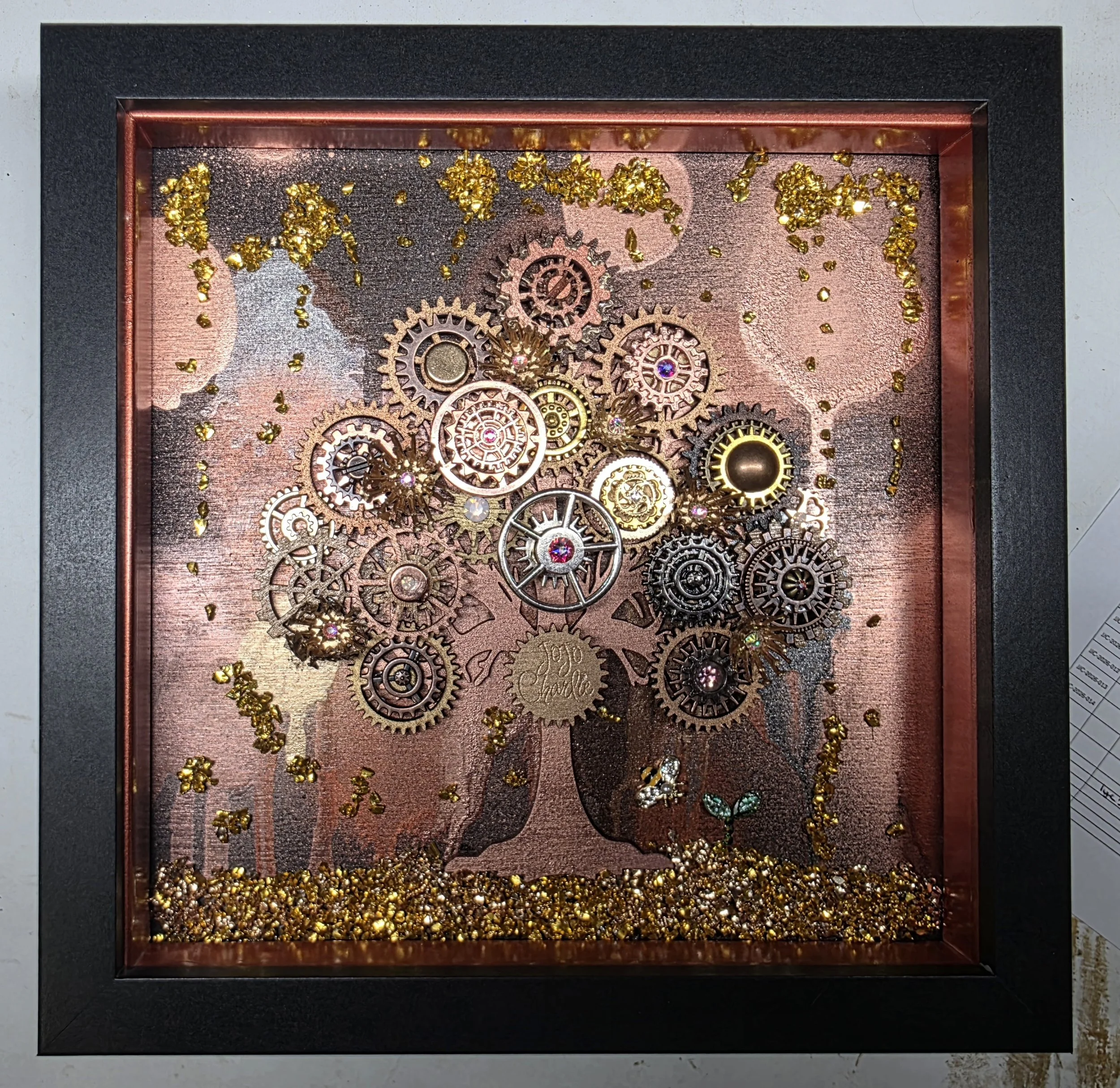 Display case with small gold decorative items, assorted cogs, and other metallic decorations on a rose gold background, surrounded by a black frame.