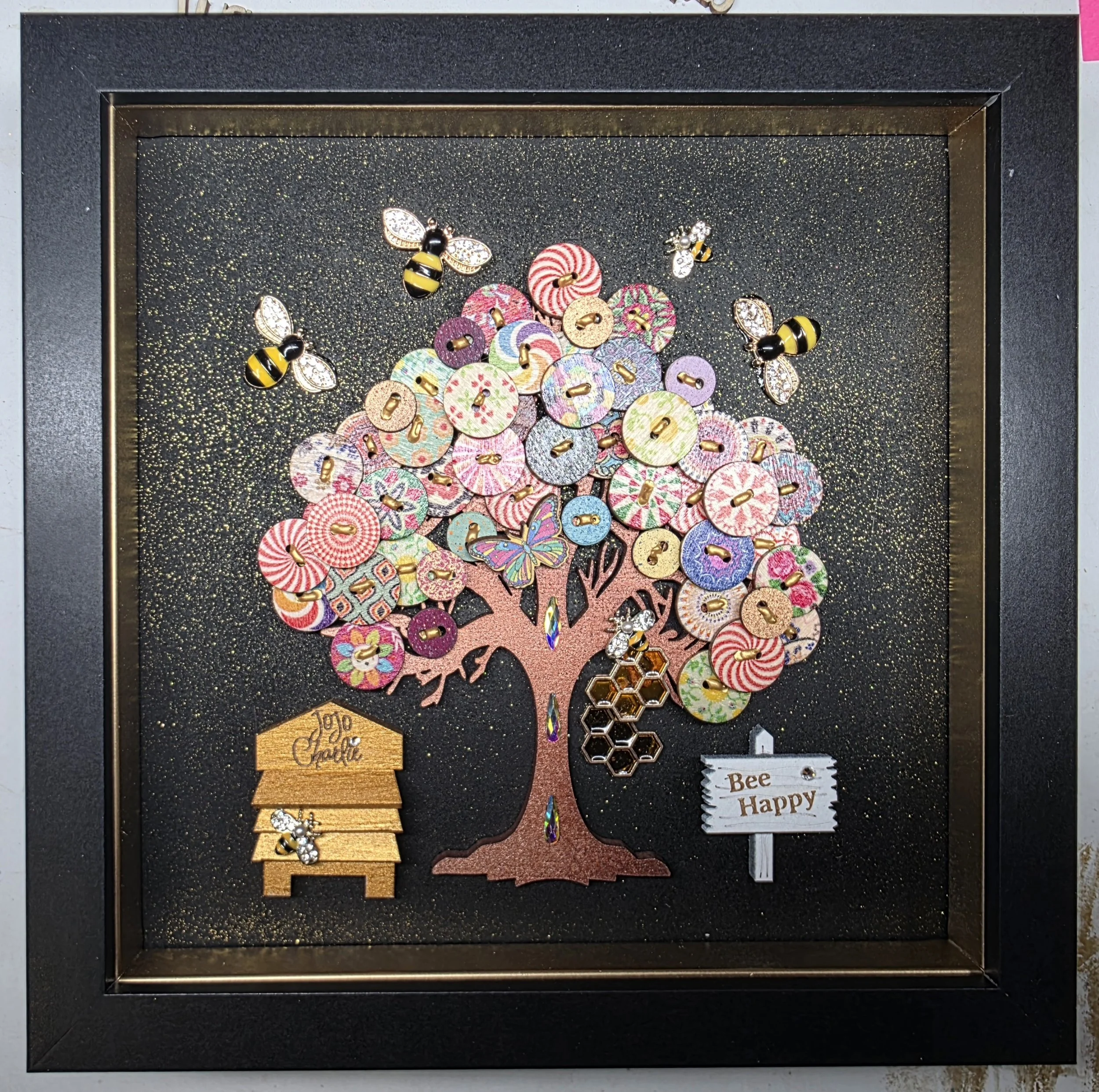 A framed display with a black background and gold glitter, featuring a decorative tree made of pink buttons with floral and swirl patterns, adorned with bee-shaped jewelry with black and yellow stripes and rhinestone wings, along with small hexagon h