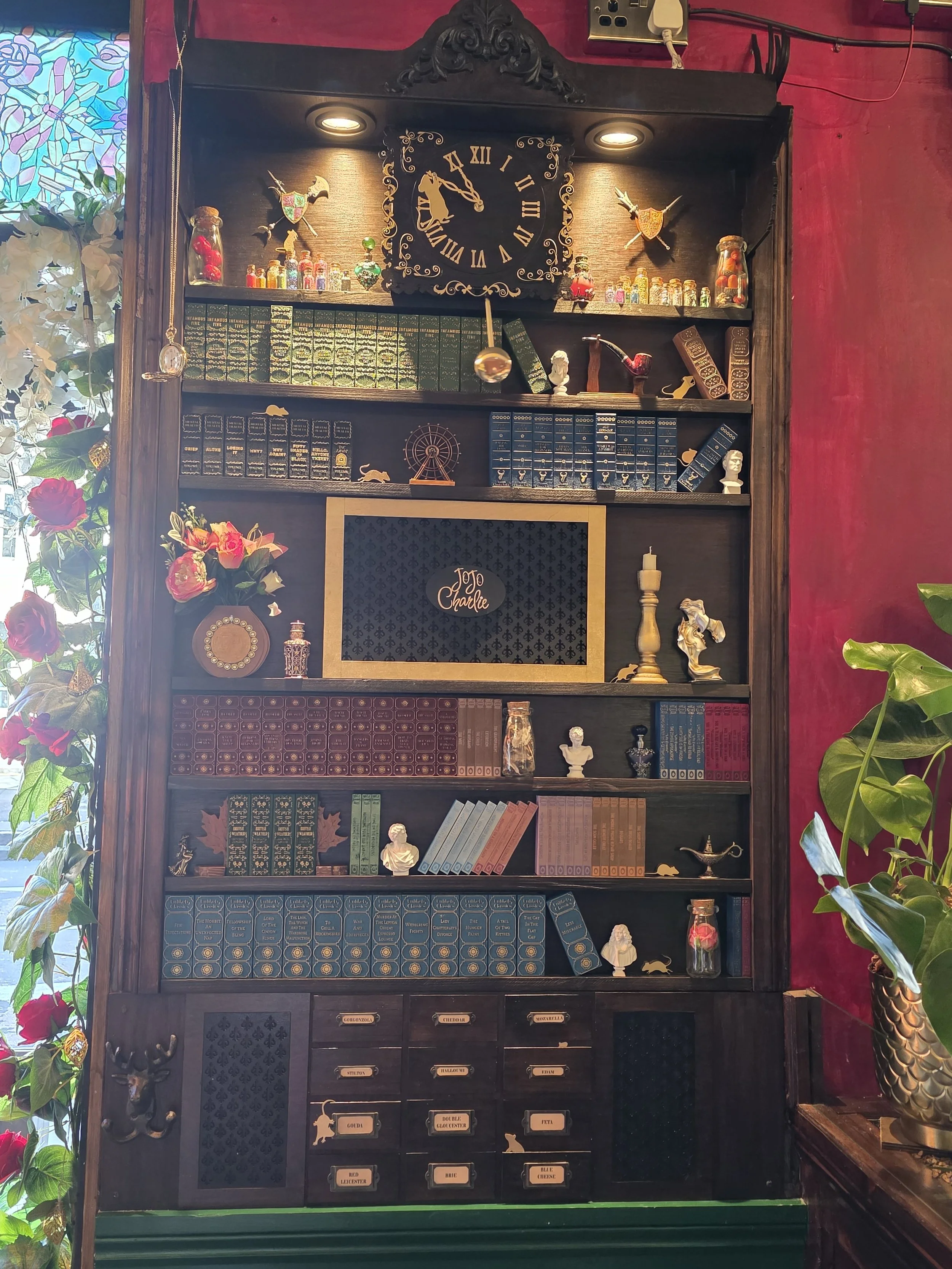 A large wooden display cabinet filled with books, curiosities, and small decorative items including busts, animals, and figurines. A clock hangs at the top center. The cabinet has labeled drawers and compartments, with plants on the sides and a stain