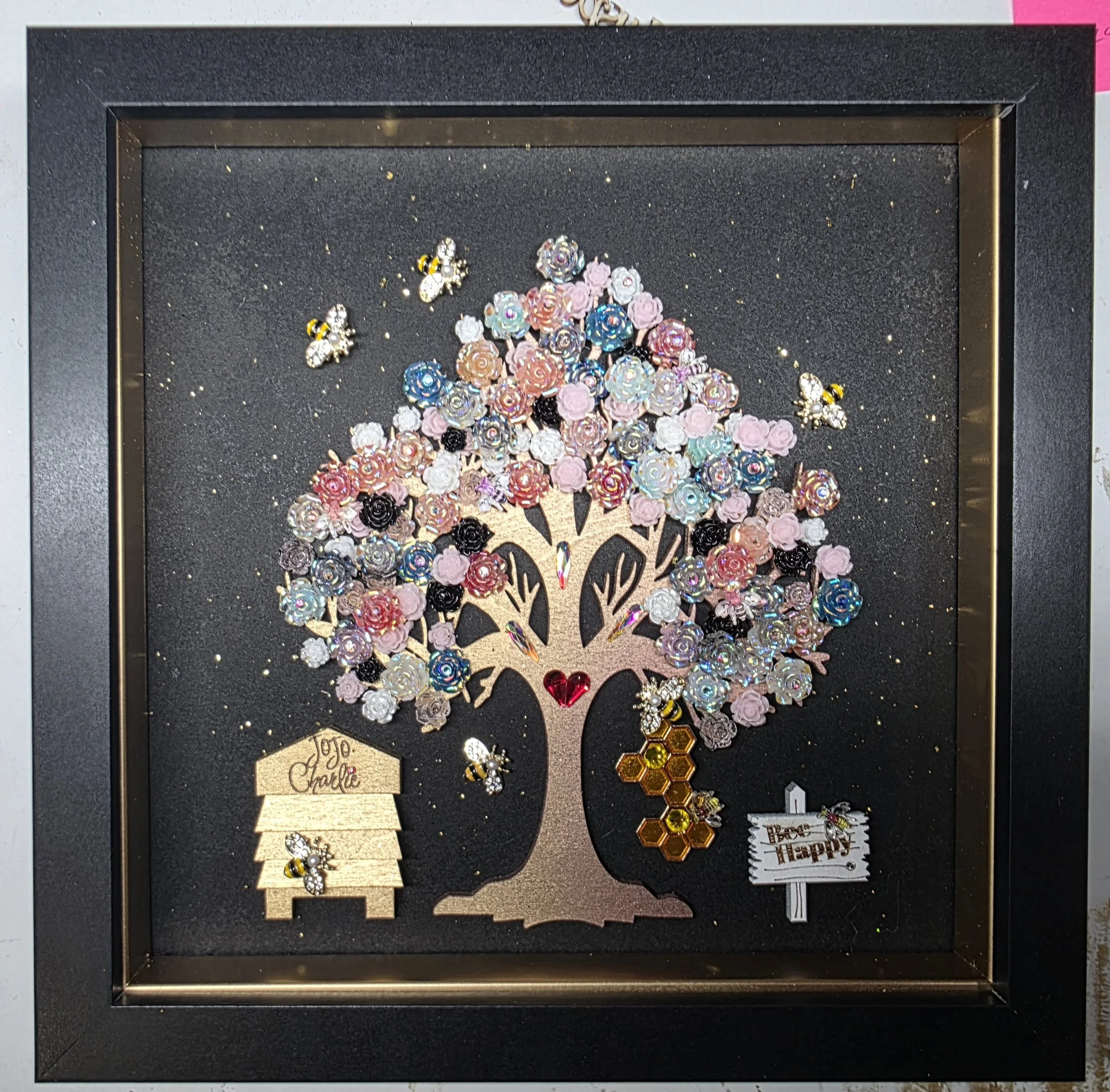 Display case with various colorful rhinestone and metal jewelry pieces, including bee, flower, and honeycomb designs on a black background with a decorative tree cutout and a small tag reading "Happy".