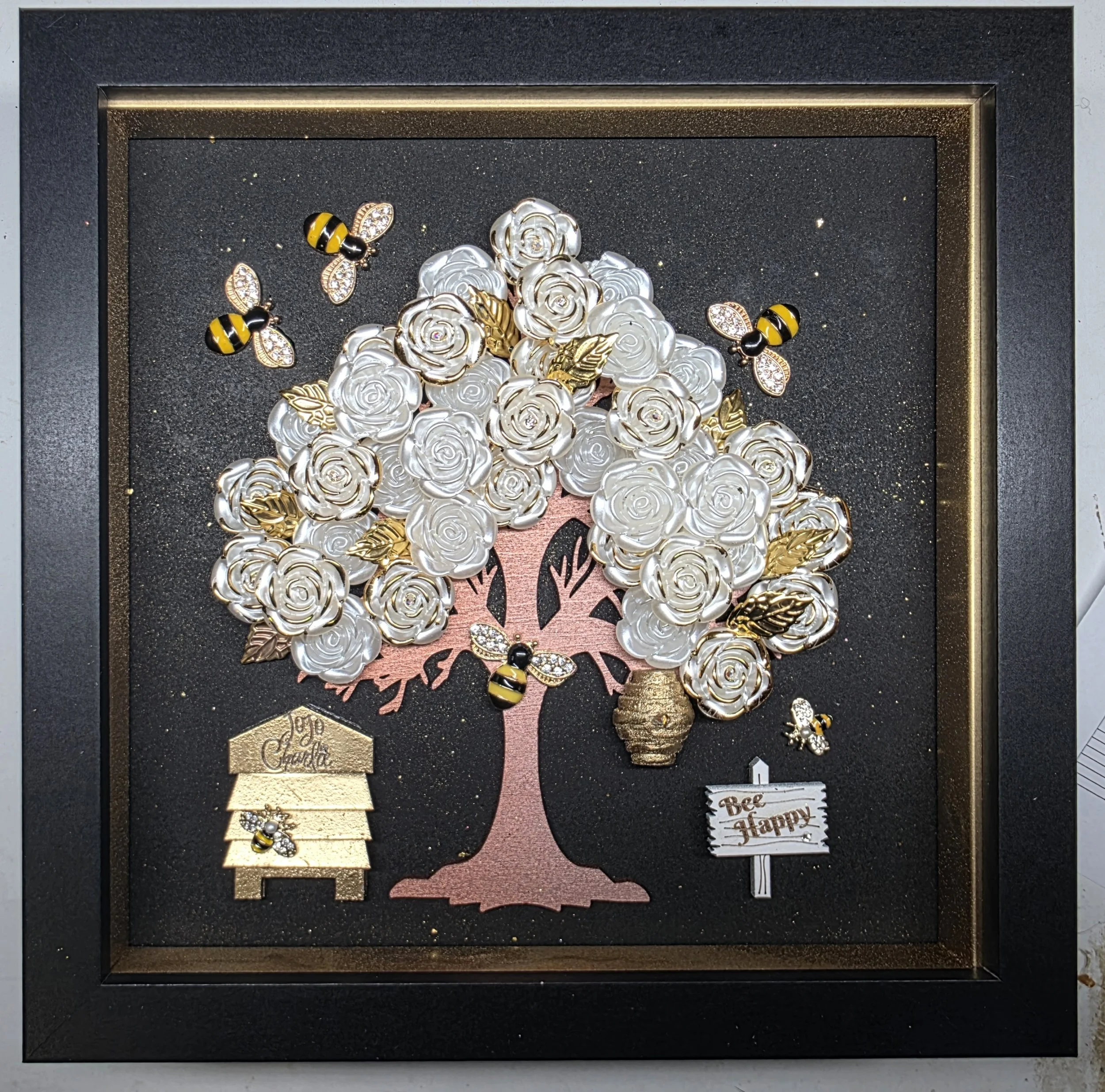 A framed decorative display featuring a bouquet of white roses made of metal or ceramic, gold leaves, and bee and leaf embellishments. There are small signs that say "joy" and "be happy" and the background has a black glittery surface.