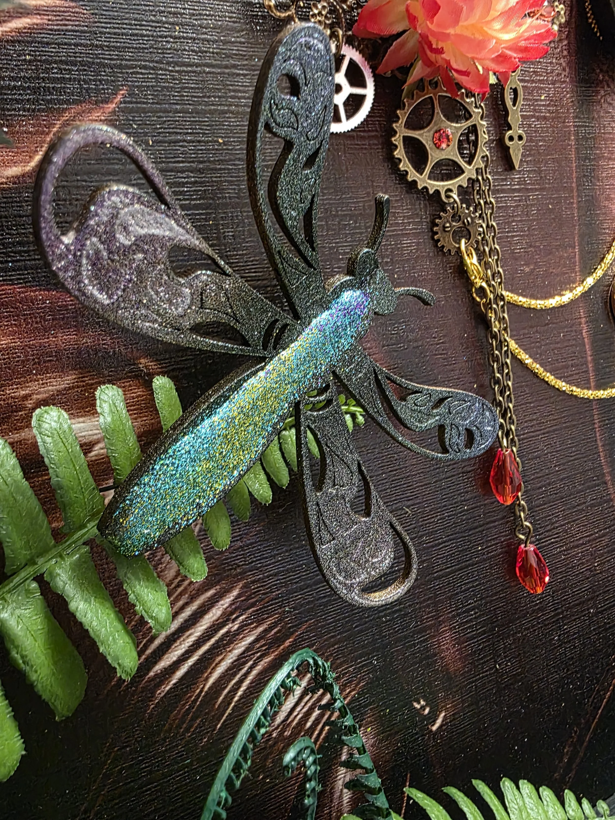A decorative butterfly jewelry piece with green and blue glittery wings, placed on a dark wooden surface with green fern leaves underneath. There is a cluster of colorful artificial flowers, gears, and chains with red and orange gemstones nearby.