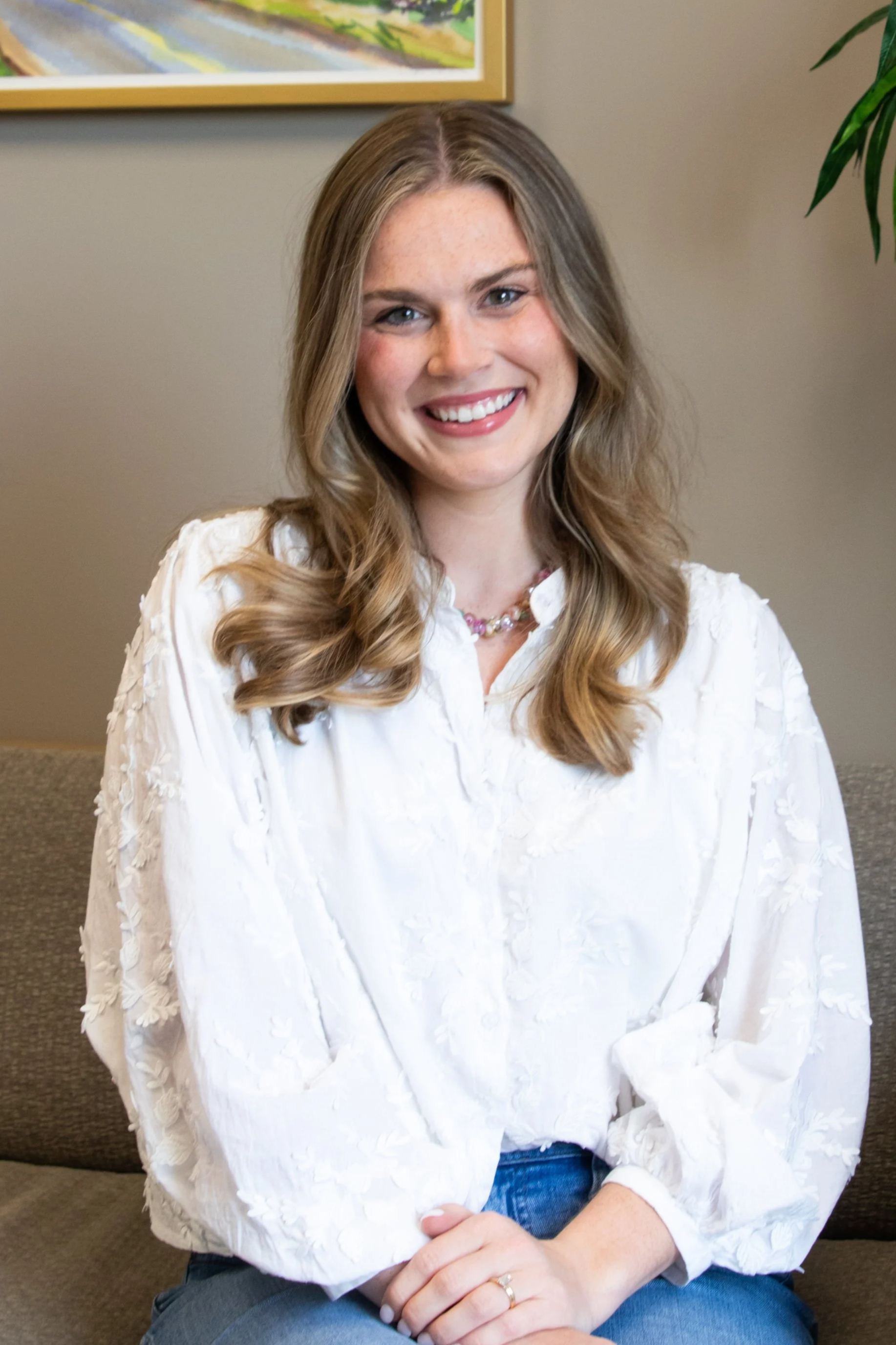 A smiling woman with long wavy light brown hair, wearing a white embroidered blouse, denim jeans, and a colorful beaded necklace, sitting on a brown couch in an indoor setting with a plant and framed artwork in the background.