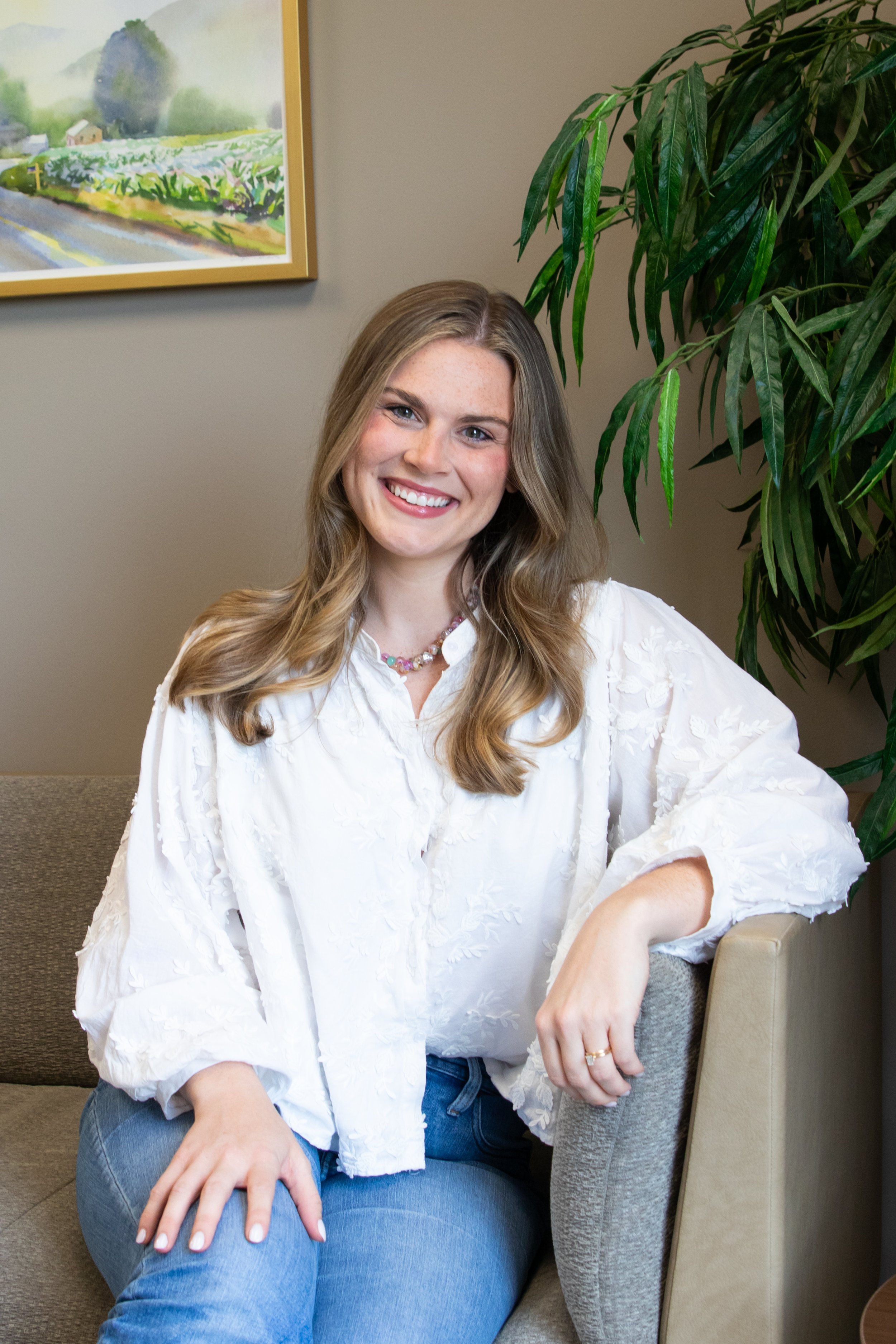 A young woman with wavy blonde hair, wearing a white embroidered blouse and blue jeans, sitting on a beige sofa next to a large green plant and a framed landscape painting on a beige wall, smiling at the camera.