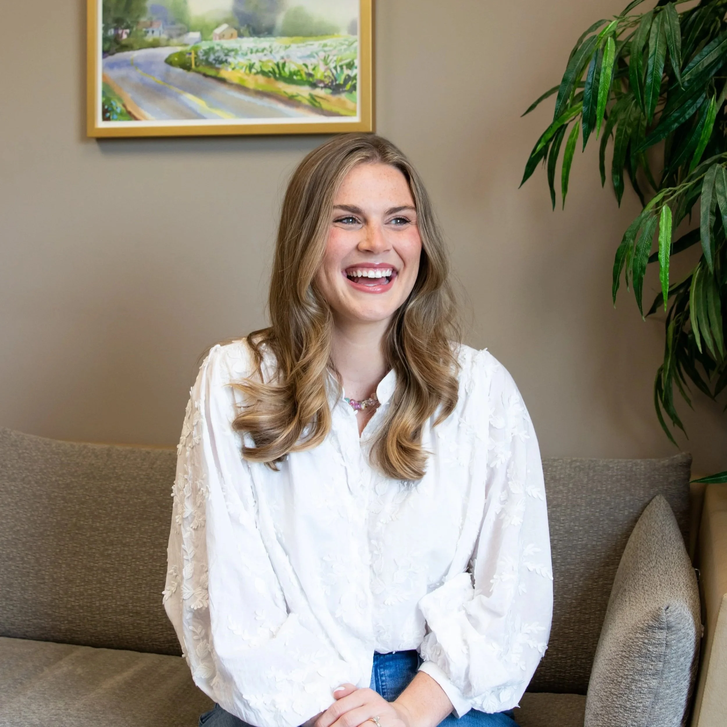 A woman with long blonde hair smiles while sitting on a gray sofa in a room with beige walls. A framed painting of a rural landscape with a road, green fields, and trees hangs on the wall behind her. To her right, there is a tall green leafy plant.