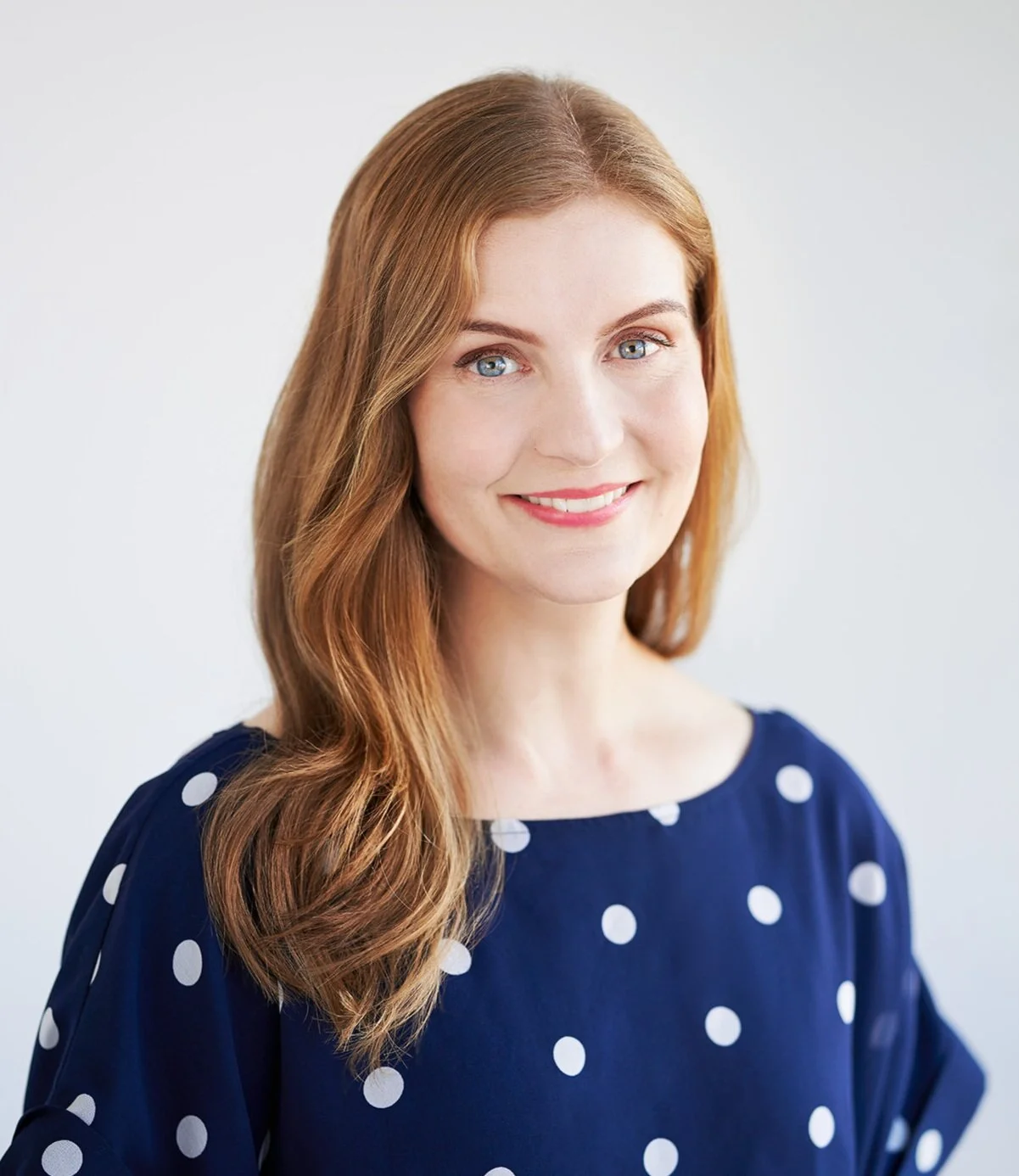A woman with long hair and blue eyes wearing a navy blue polka dot top, smiling against a plain light background.