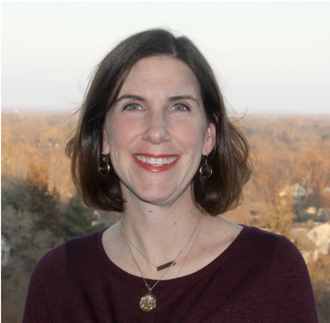 A woman with dark brown hair and blue eyes, smiling, standing outdoors with trees and houses in the background during autumn.