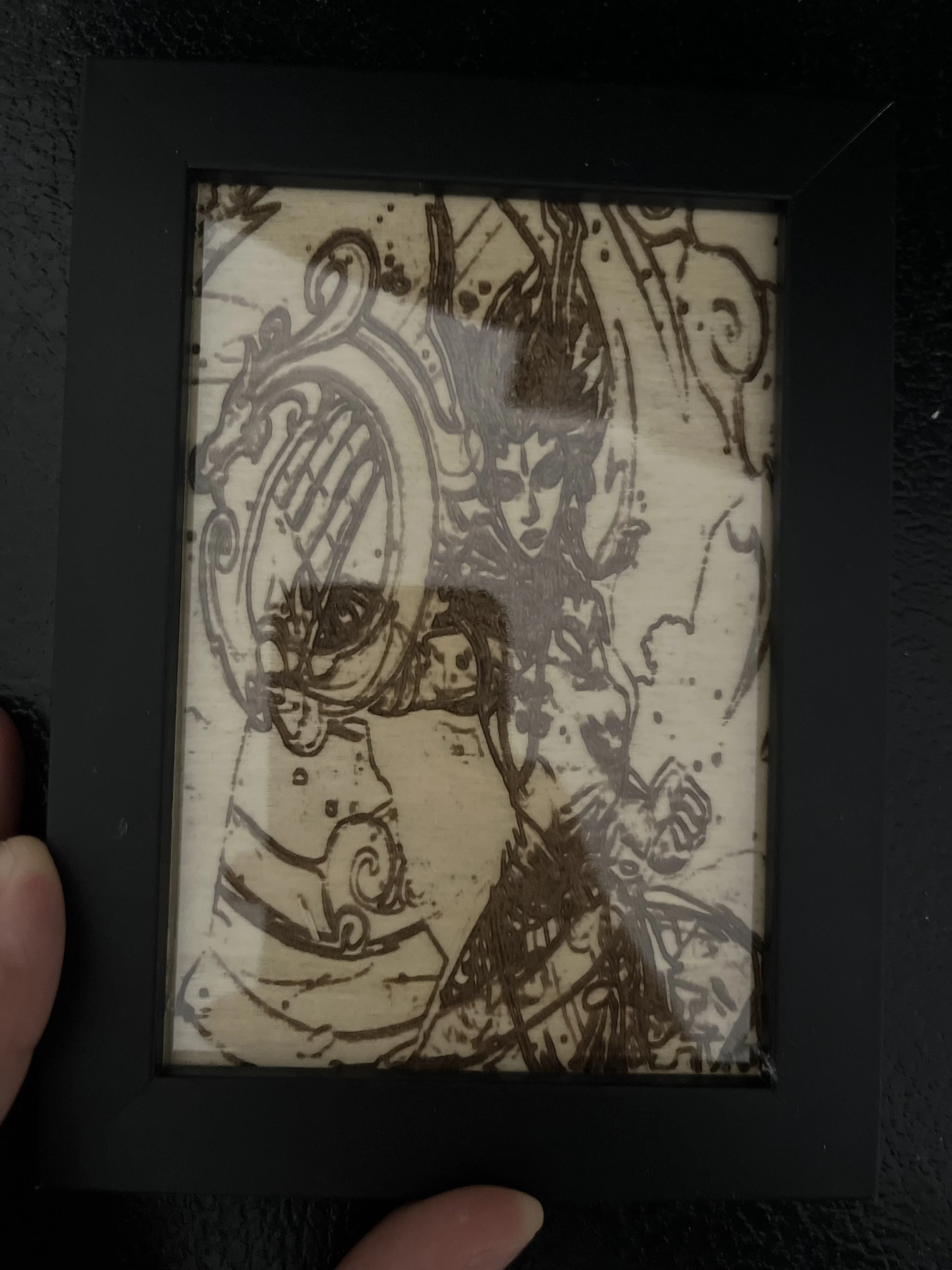 A hand holding a framed artwork featuring a line drawing of a woman with flowing hair and ornate details, printed on a light-colored background.