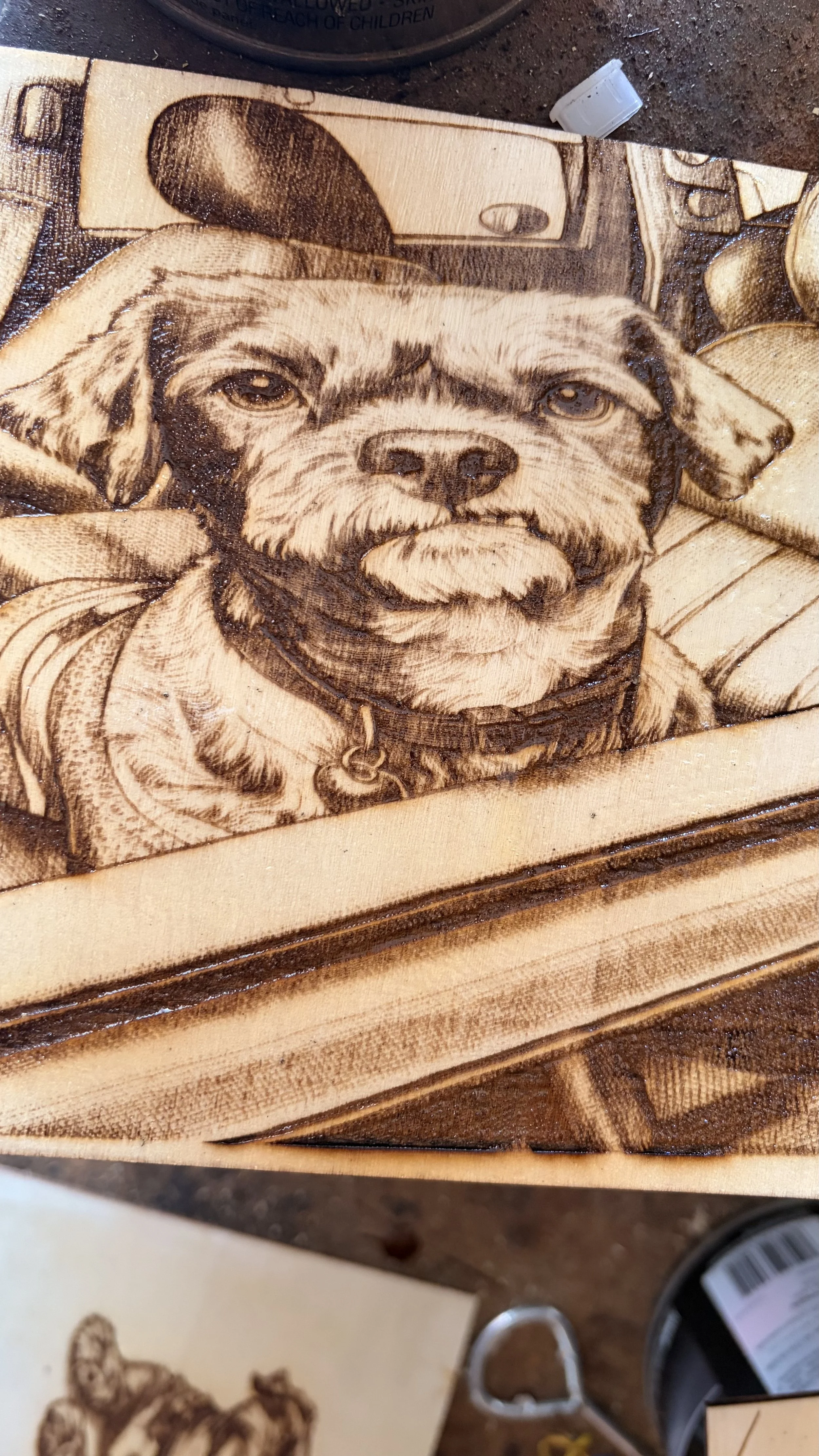 Wooden plaque with a detailed pyrographic illustration of a dog's face, wearing a collar, with a background of a landscape scene, and some tools and items on a nearby table.