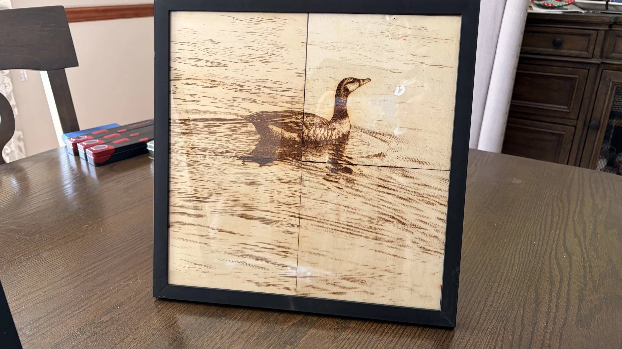 Framed wooden picture of a mallard duck swimming on water on a table, with furniture and objects in the background.