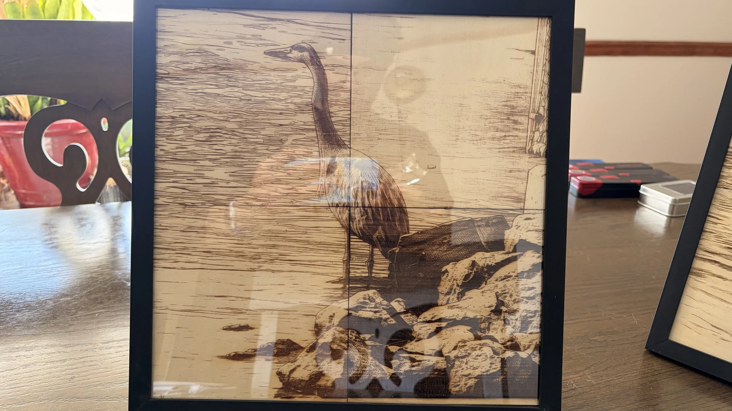 Framed artwork of a bird standing on a log, inside a black frame, placed on a wooden table.