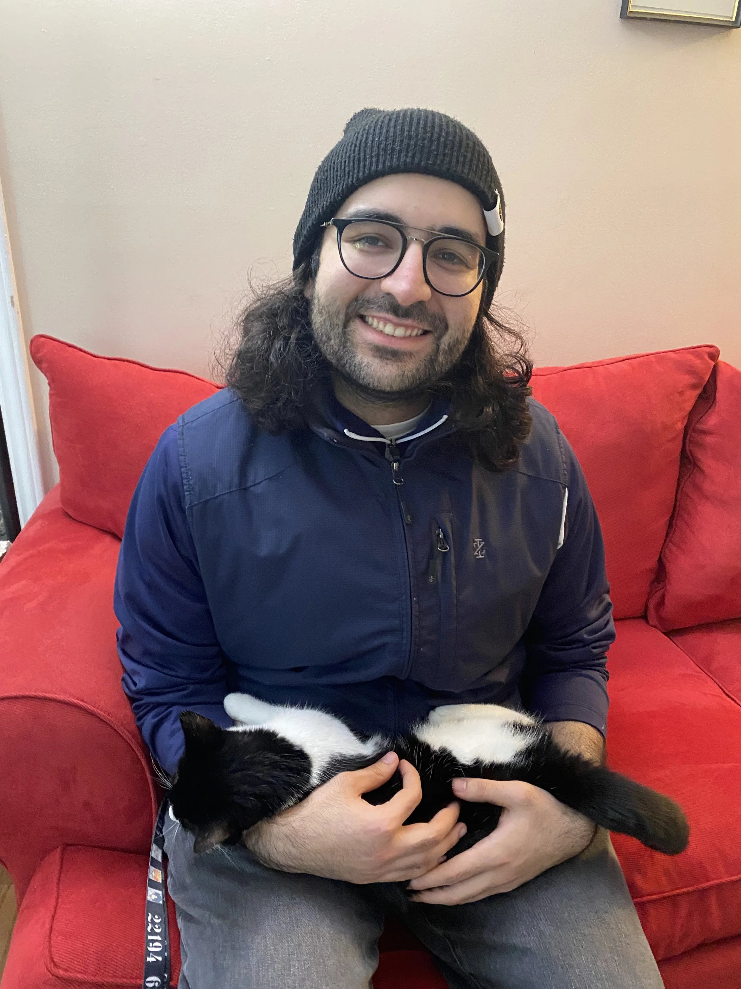 A man with long curly hair, glasses, and a black beanie sitting on a red couch, smiling and holding a black and white cat.