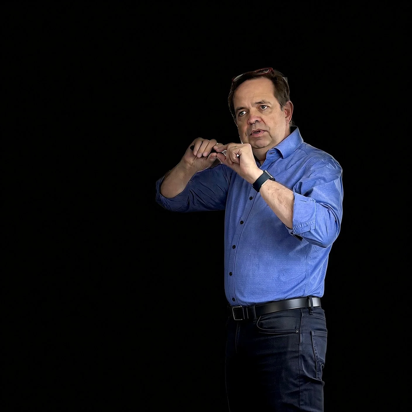 A man in a blue shirt and black pants stands against a black background, adjusting his glasses with a serious expression.