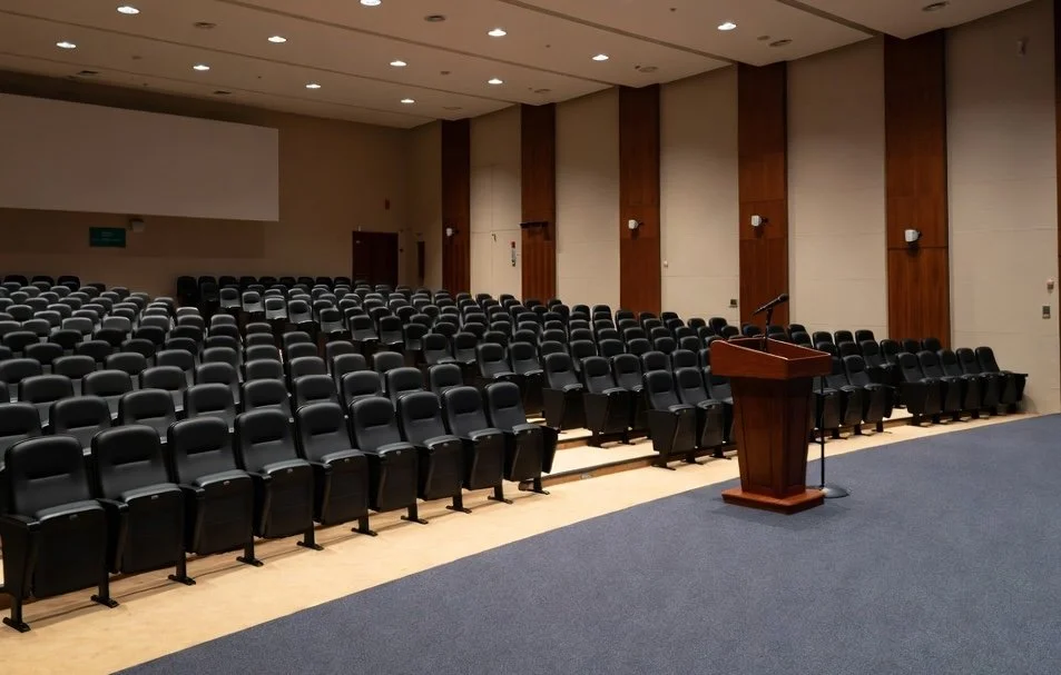Empty conference room with rows of black chairs, a wooden podium with a microphone, and a large speaker screen on the wall.