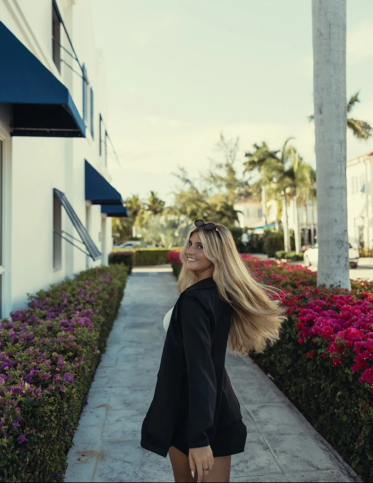 A young woman with long blonde hair standing on a sidewalk lined with purple and pink flowers, smiling and looking over her shoulder, wearing a black blazer and sunglasses.