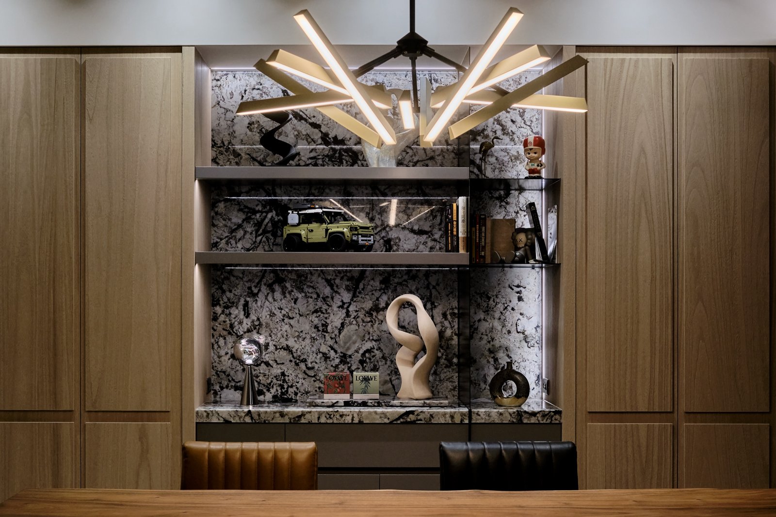 Modern built-in shelving unit with decorative items, books, and a model vehicle, set against a black and white marble wall and illuminated by a contemporary chandelier, with wooden cabinetry on both sides.