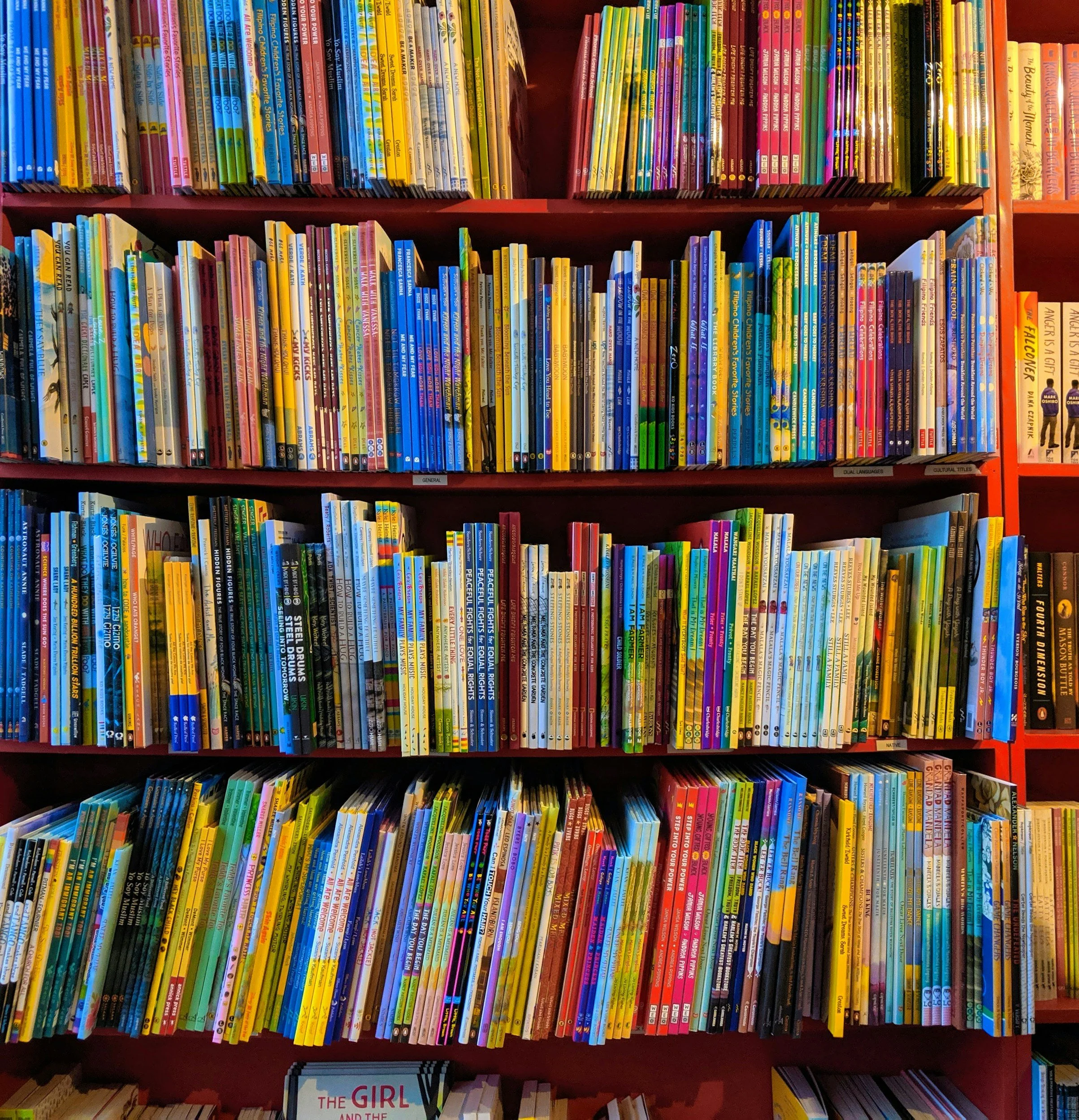 Shelf of colorful children's books arranged by color on red shelves in a bookstore or library.