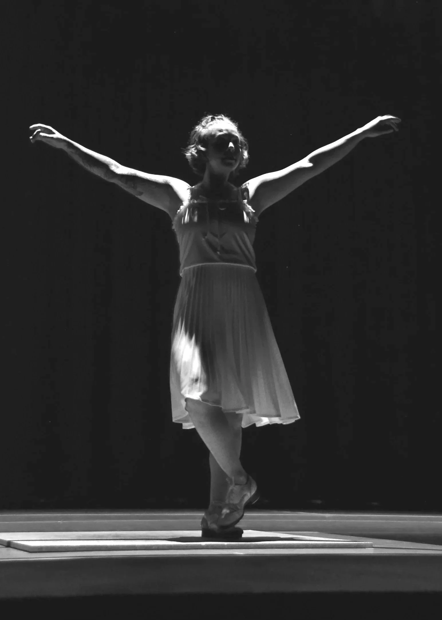 Black and white photo of a ballet dancer on stage, wearing a dress and pointe shoes, with arms outstretched and smiling.