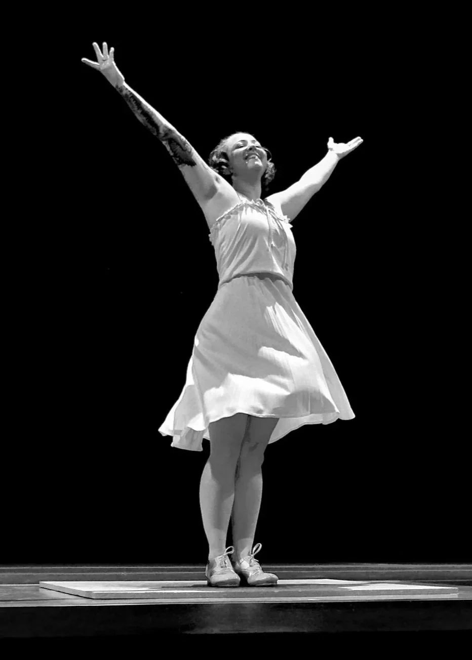 A woman standing on a stage with her arms raised and outstretched, wearing a dress and sneakers, smiling with her eyes closed.