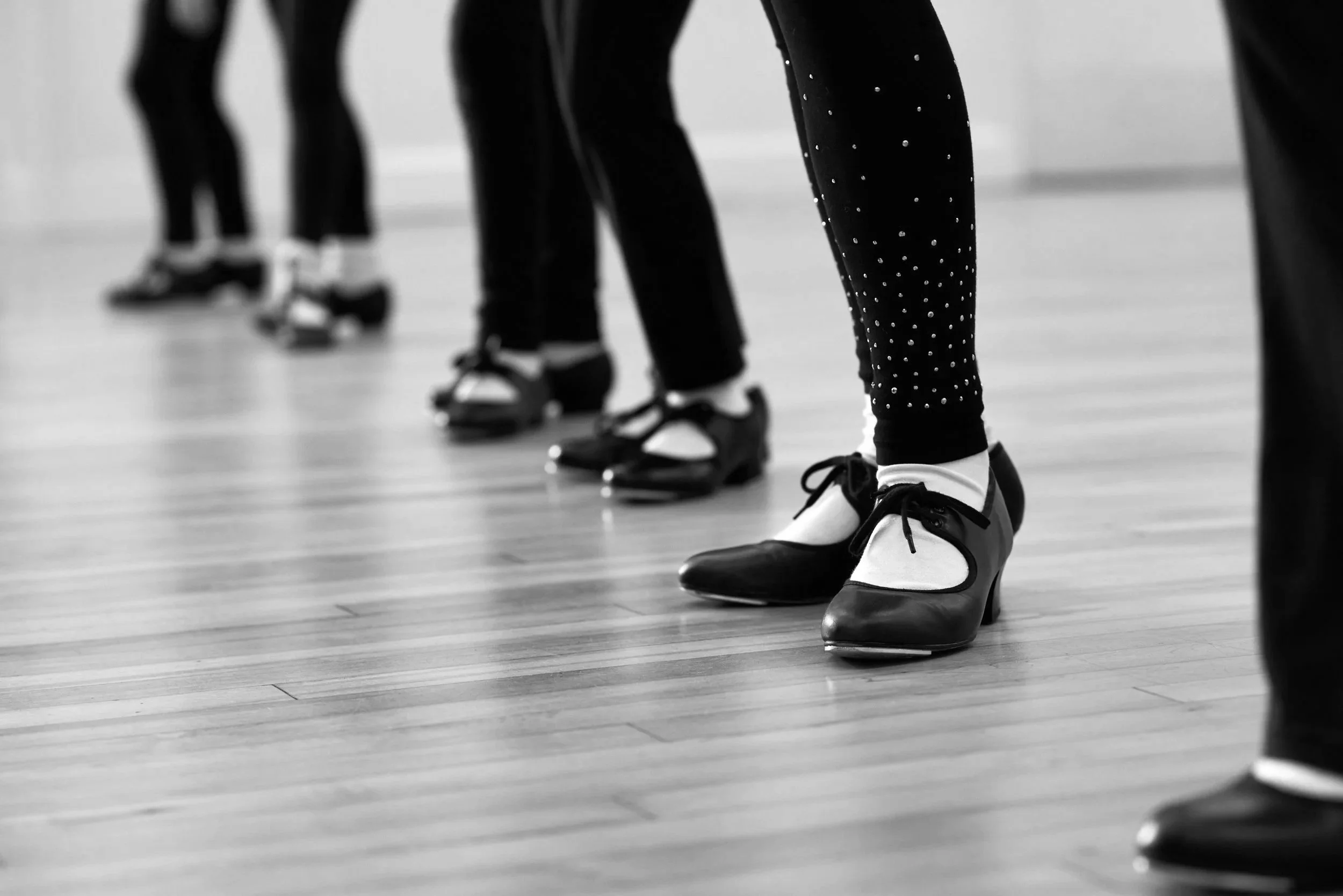 Close-up of ballet dancers' feet and legs, wearing pointe shoes and athletic clothing, standing on a wooden dance floor.