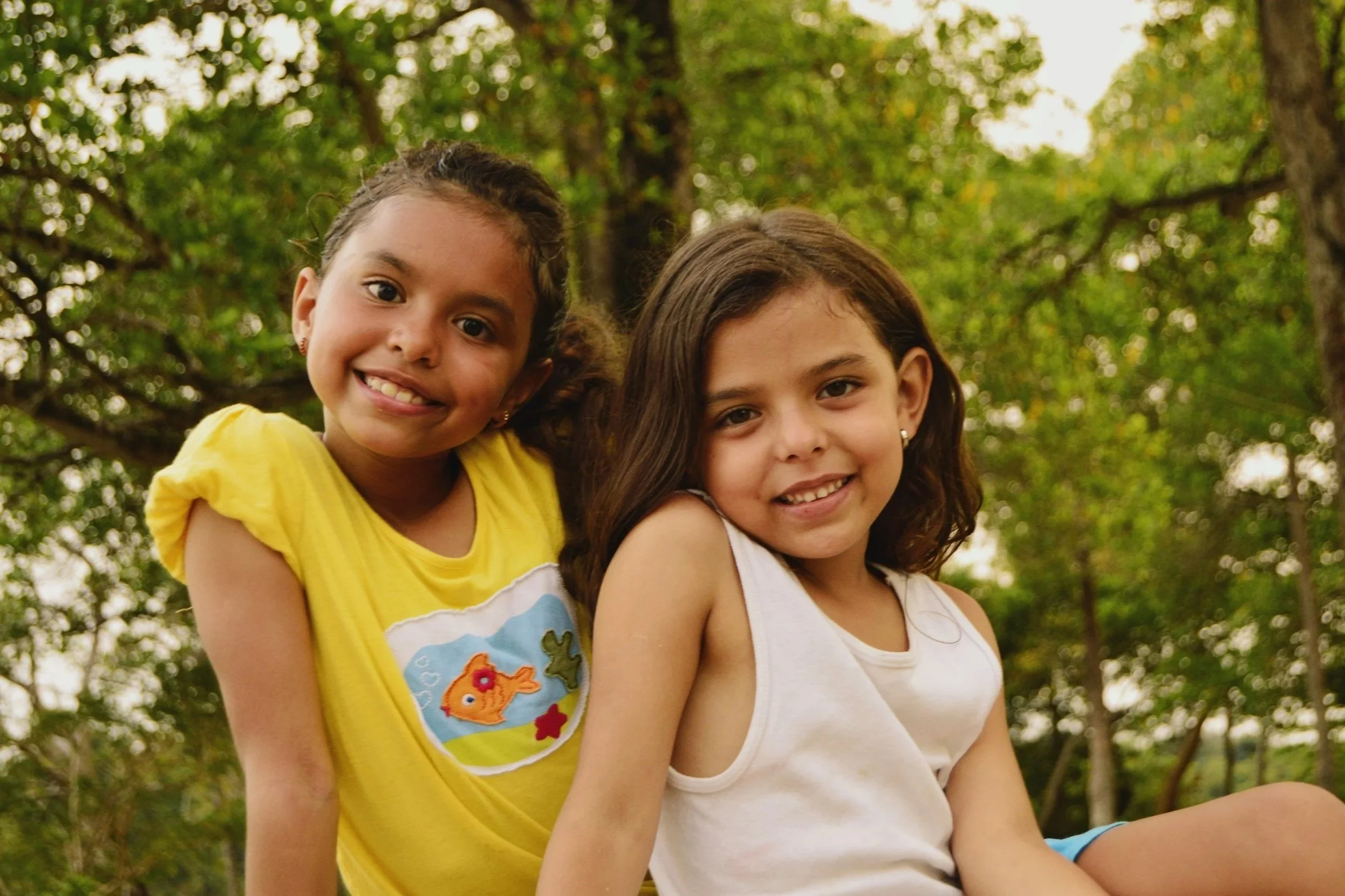 Two young girls, one with curly hair and a T-shirt with a fish design, and the other with straight brown hair and a white tank top, smile outdoors in a green, leafy setting.