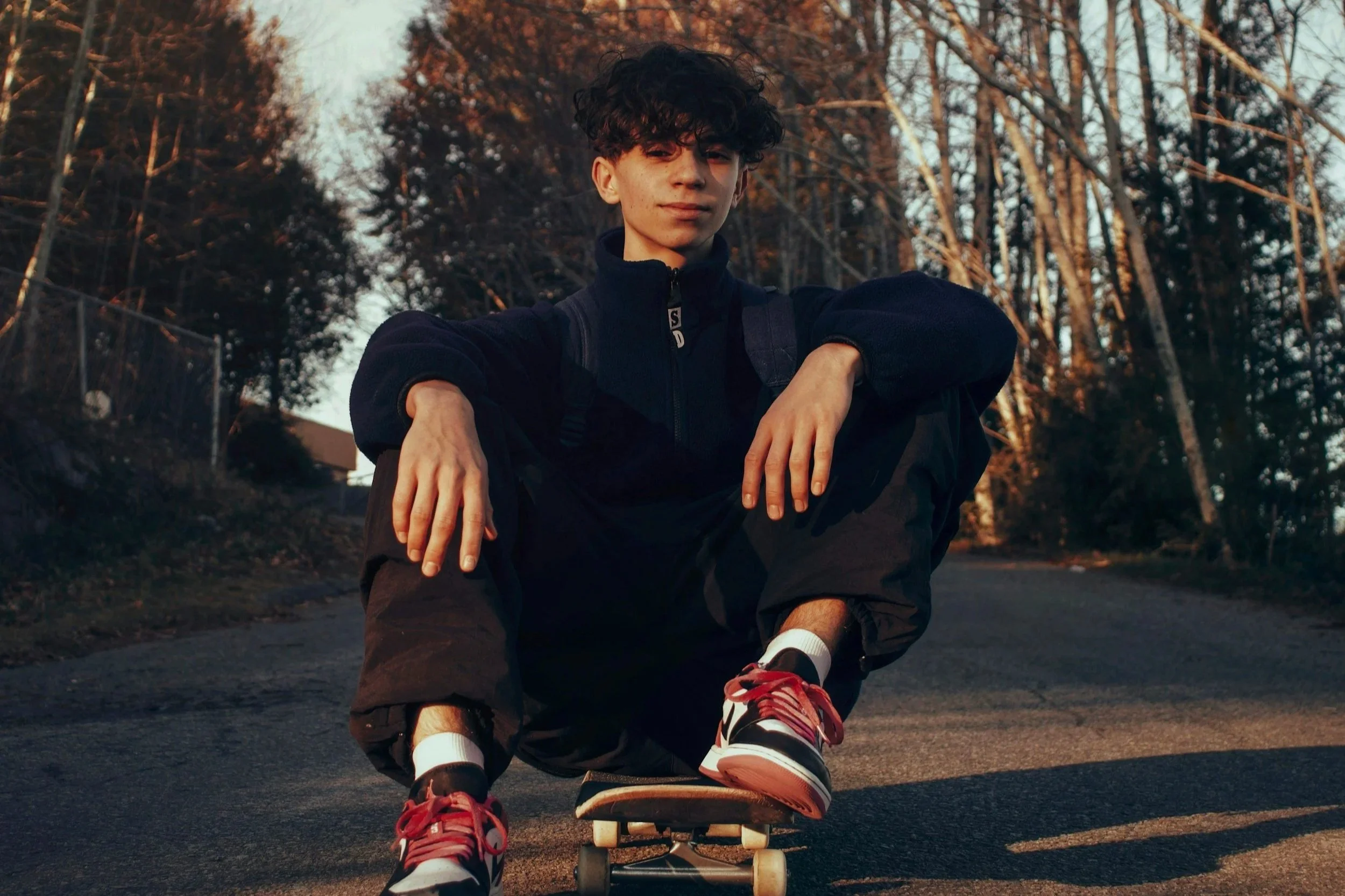 A young man with curly hair, wearing a dark jacket and backpack, skateboarding outdoors on a paved surface during sunset with trees in the background. Teenager with anxiety, SEMH, mental health