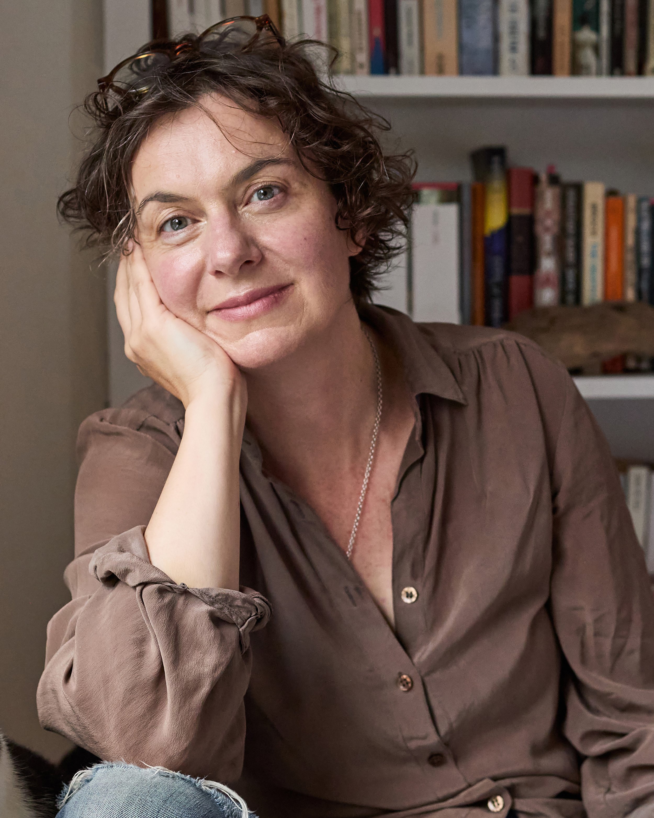 Portrait of author Tiffany Watt Smith, a woman with short, curly brown hair resting her face on her hand, sitting in a room with a bookshelf in the background.