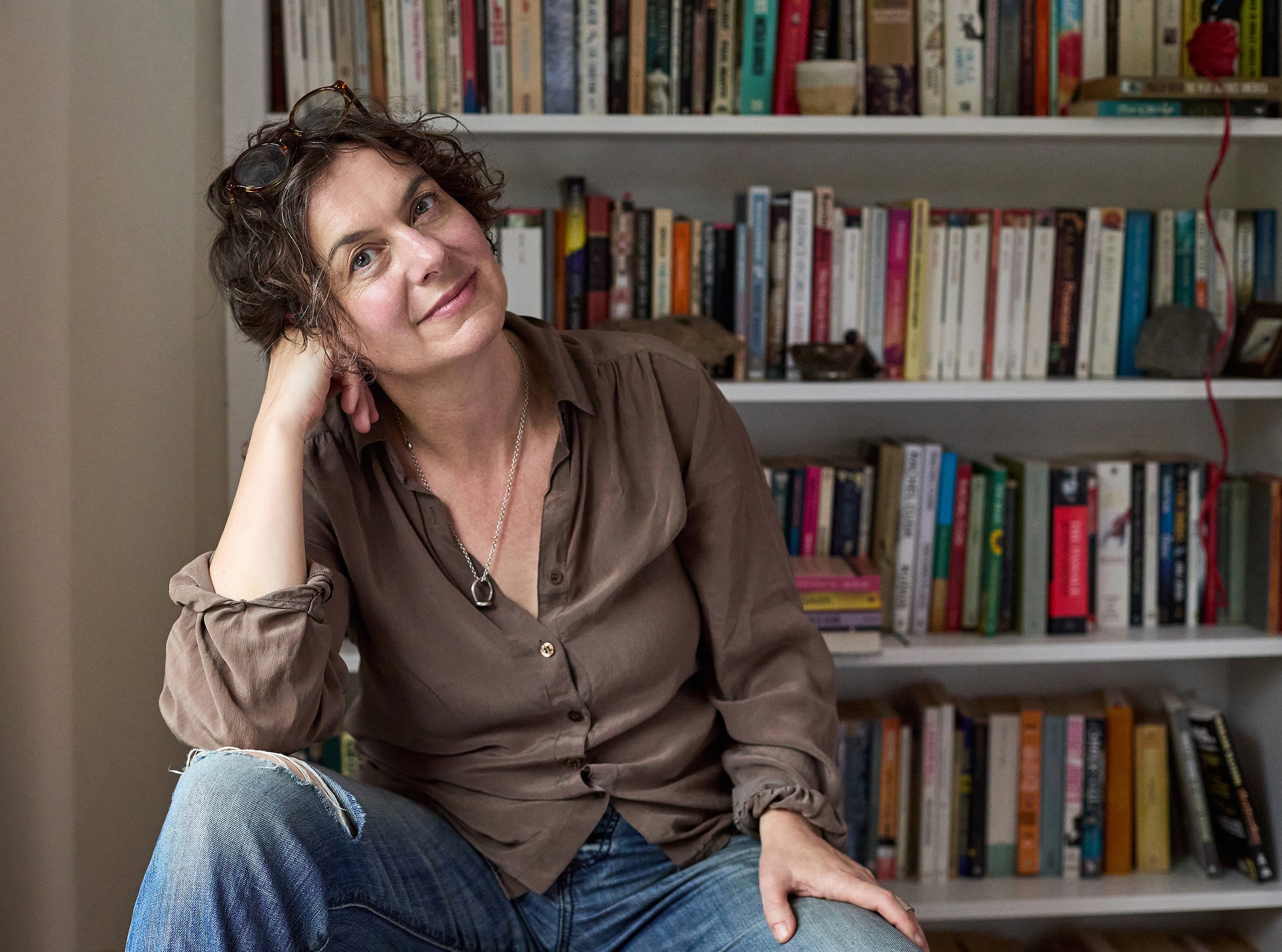 Portrait of Author Tiffany Watt Smith, woman with short brown curly hair wearing jeans with a bookcase behind her