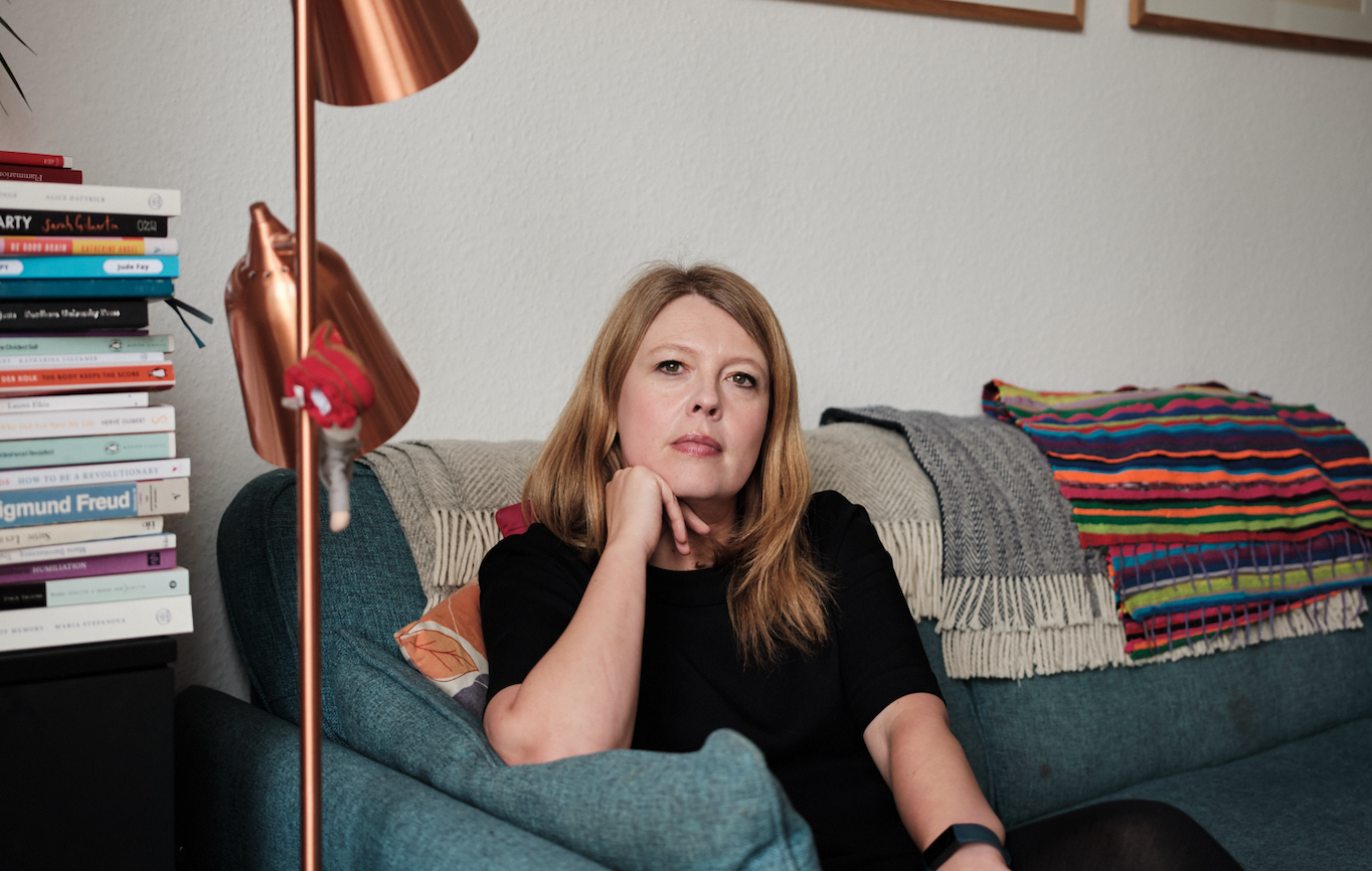 A woman with shoulder-length reddish blond hair sitting on a blue-green sofa, resting her chin on her hand, looking attentively. Behind her are stacked books, and colorful blankets are draped over the back of the sofa. To her left is a copper-colored lamp.