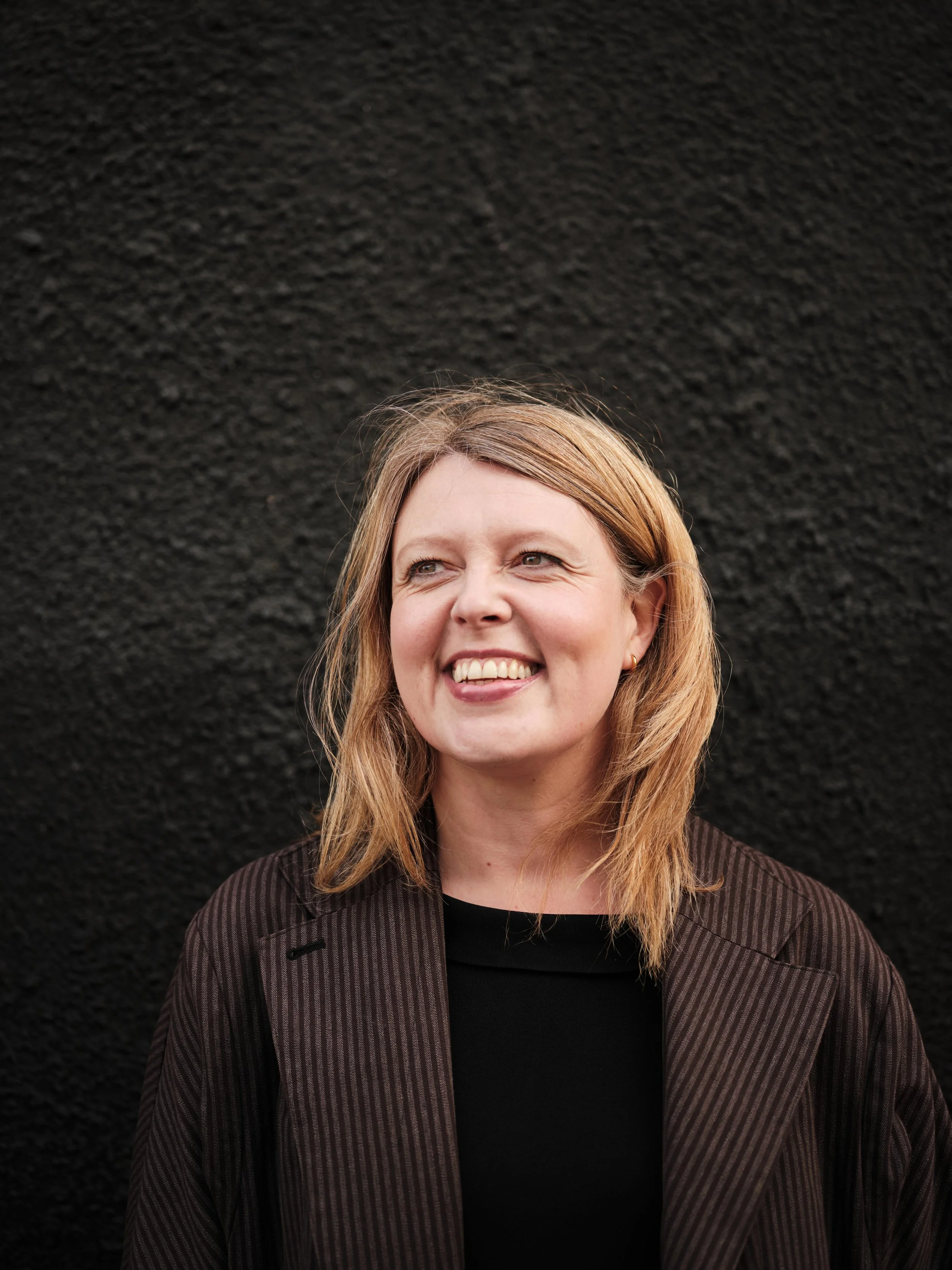 Portrait of author Katherine Angel, a woman with blonde hair wearing a dark striped blazer and a black shirt, smiling against a black textured background.