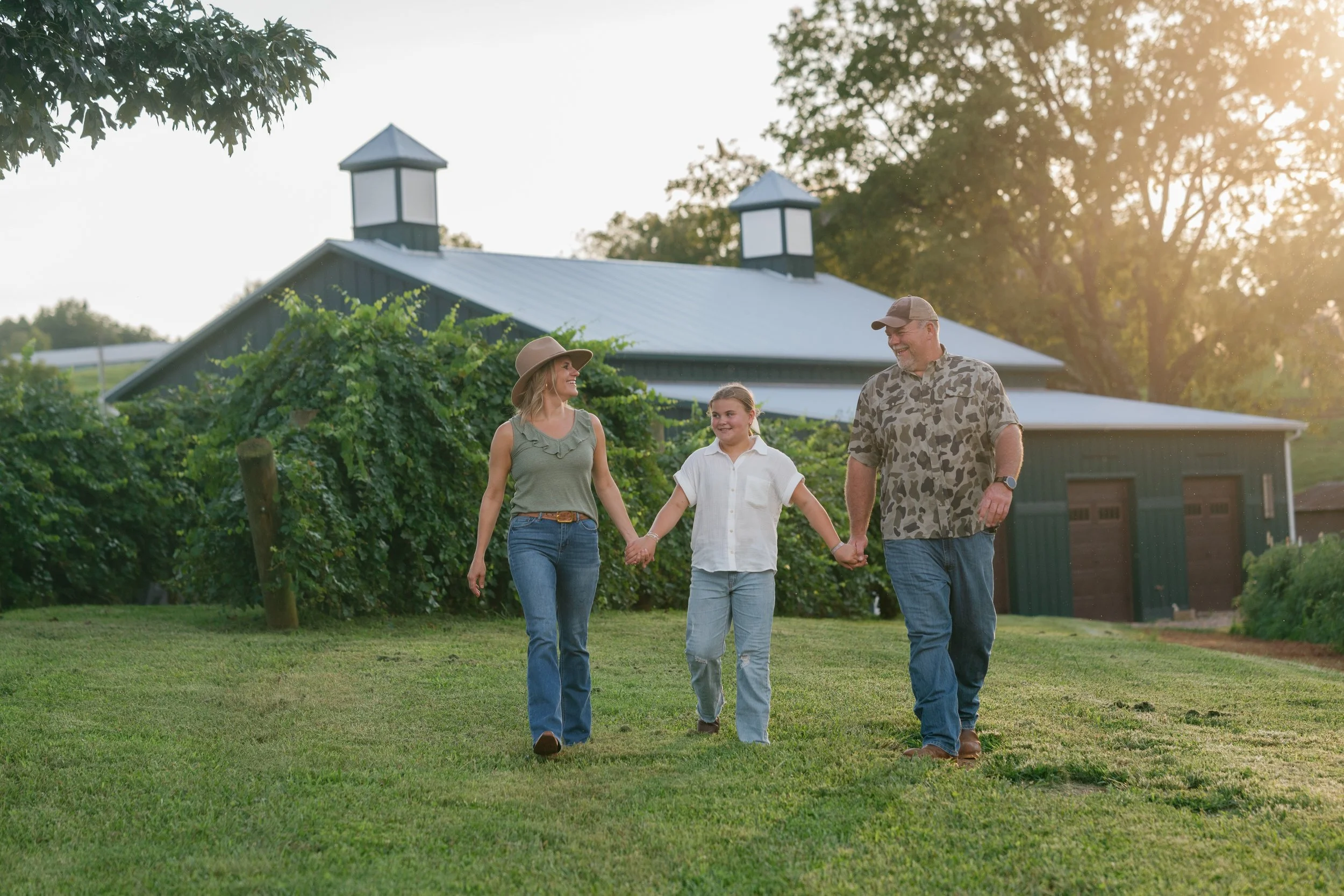 The Hulsey family,  walking hand in hand across Bright Meadows Farm with trees and a barn in the background during sunset.
