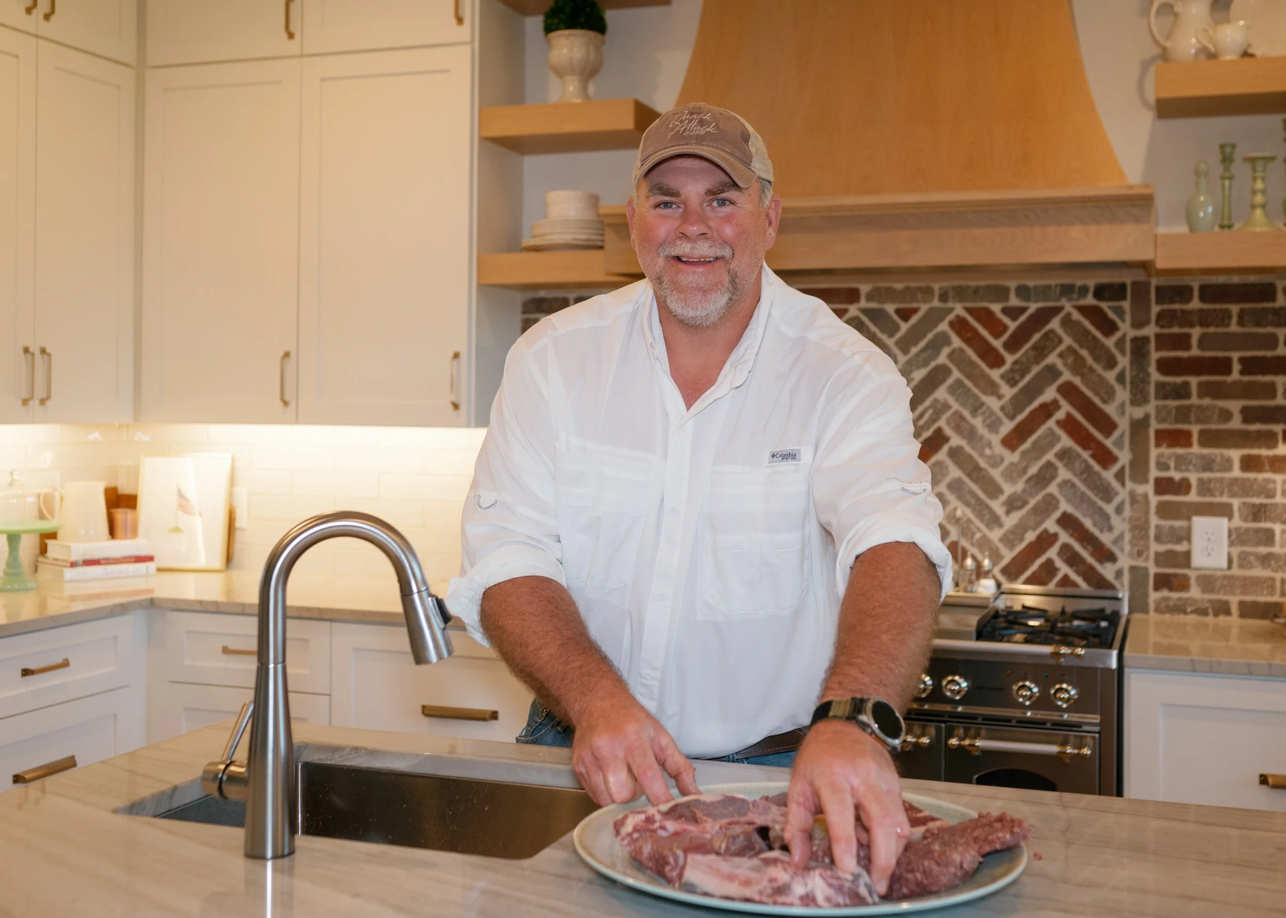 Corey Hulsey in a white shirt and beige cap is preparing Bright Meadows Farm Japanese wagyu on a kitchen countertop. The kitchen has white cabinets, a brick and wooden backsplash, and shelves with decorative items and plates.