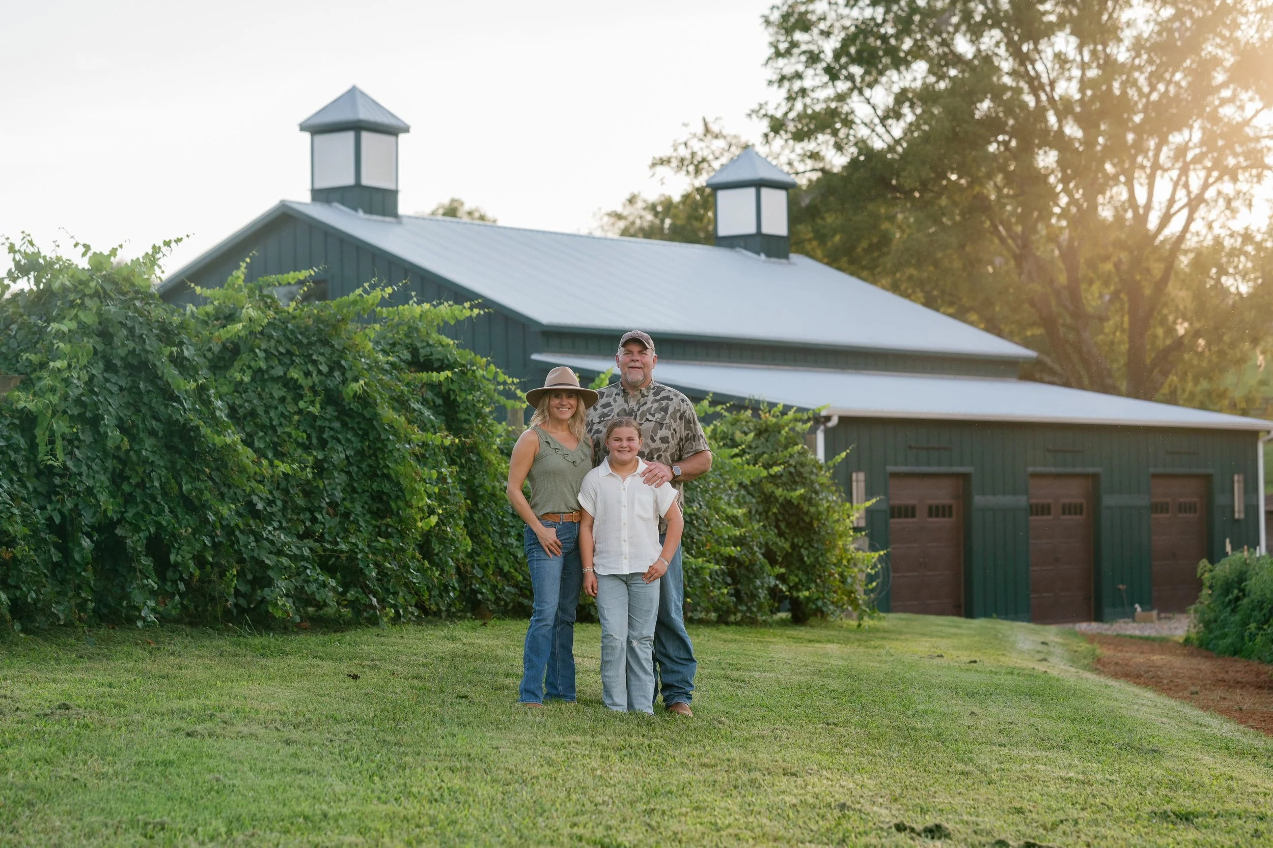 The Hulsey family, standing outdoors in a grassy yard in front of a green barn on Bright Meadows Farm with a metal roof, surrounded by trees, with sunlight shining from the right.