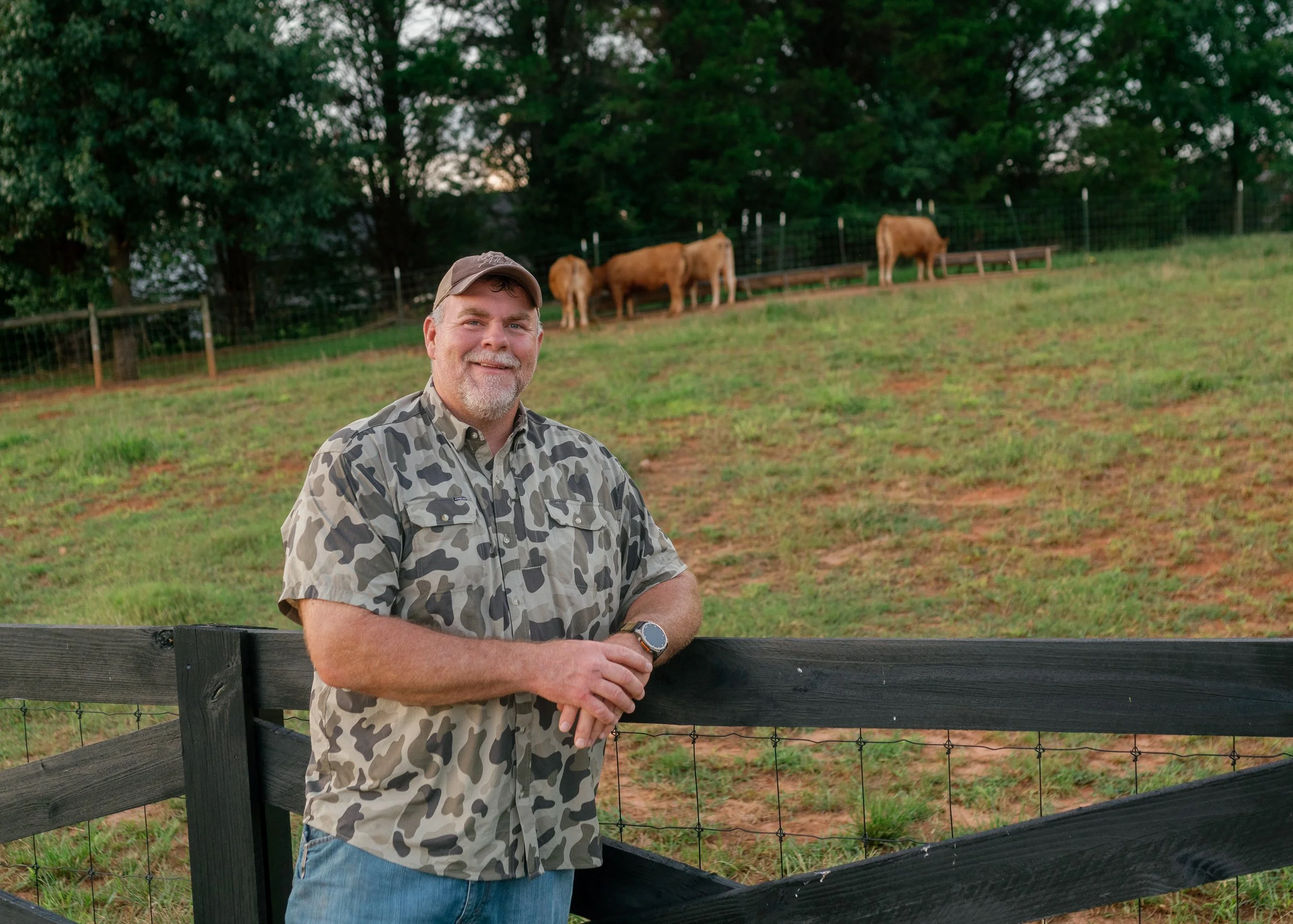 Corey Hulsey, smiling, wearing a camouflage shirt and a cap, leaning on a black wooden fence, with a field of grass, trees, and three brown wagyu cows in the background.