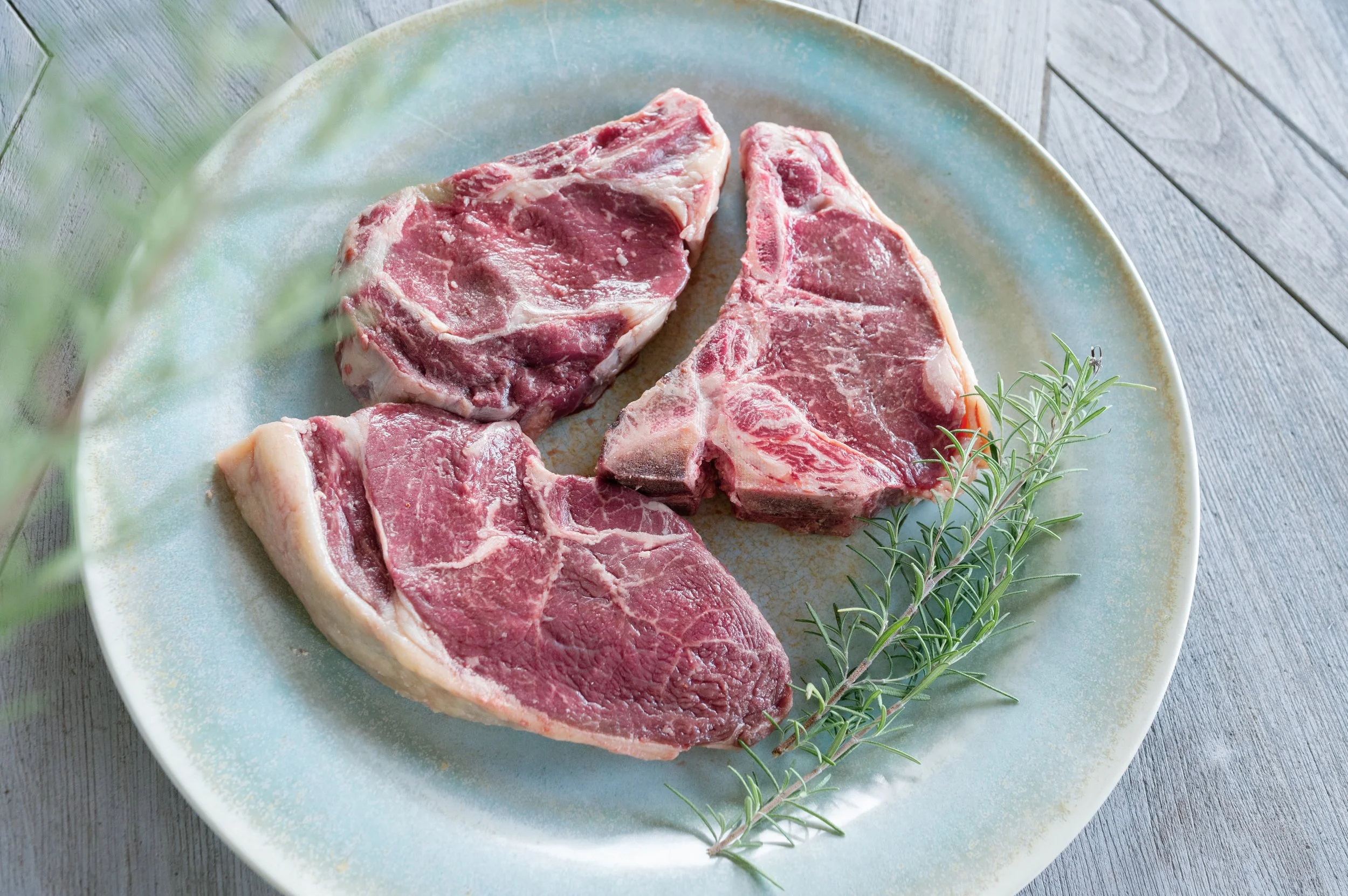 Three raw wagyu T-bone steaks on a white plate with a sprig of rosemary, placed on a wooden surface.