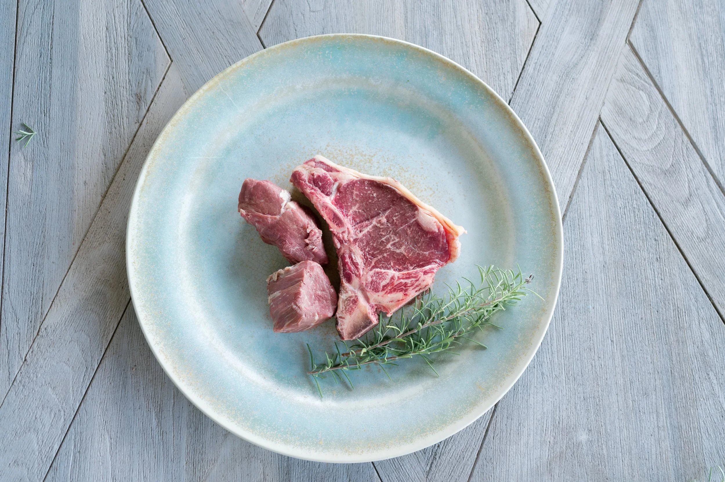 Raw wagyu beef on a blue ceramic plate with sprigs of herbs on a wooden surface.