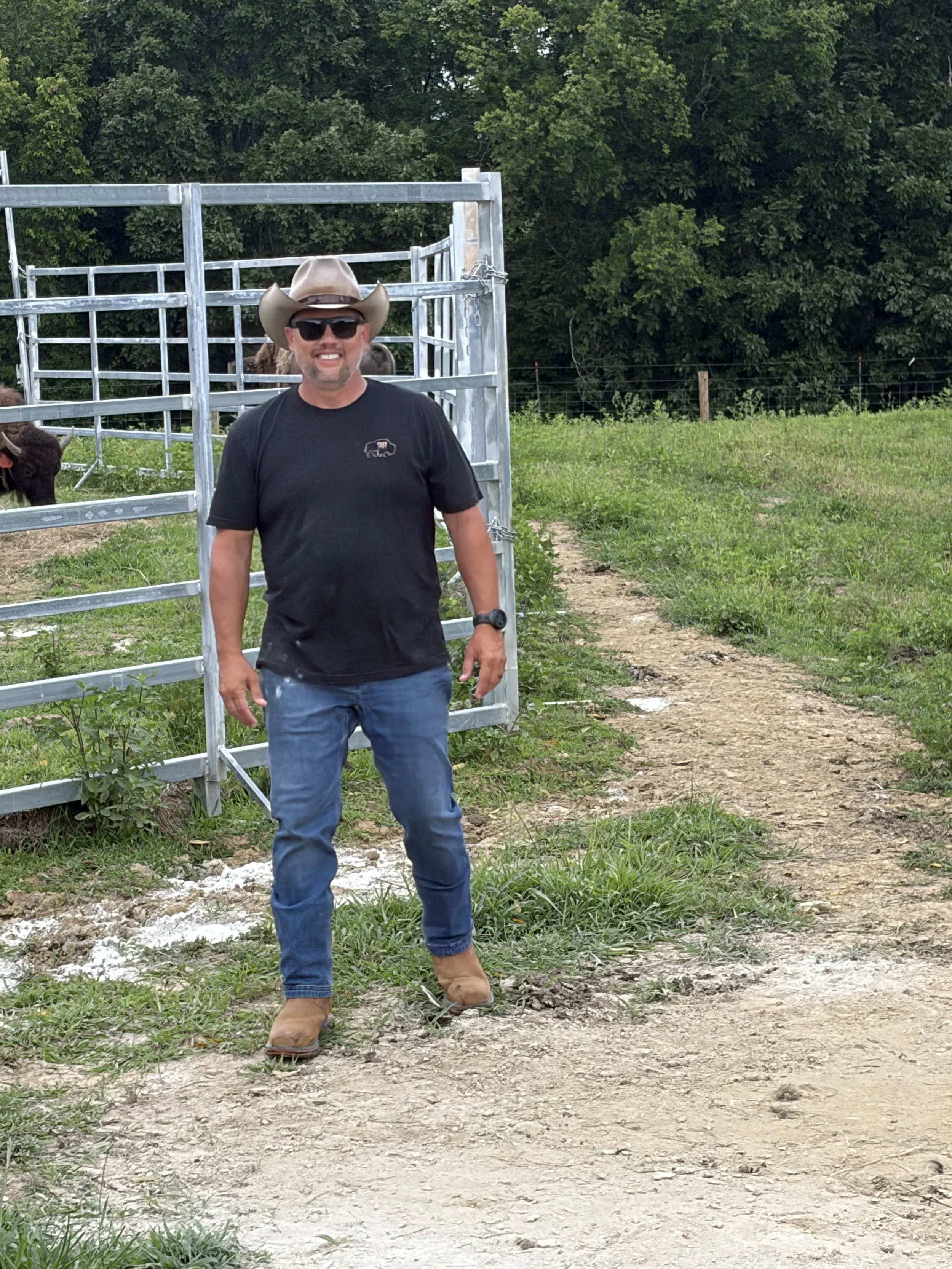A man wearing a tan cowboy hat, black sunglasses, black T-shirt, and jeans, standing outside on dirt ground near a metal gate with green grass and trees in the background.