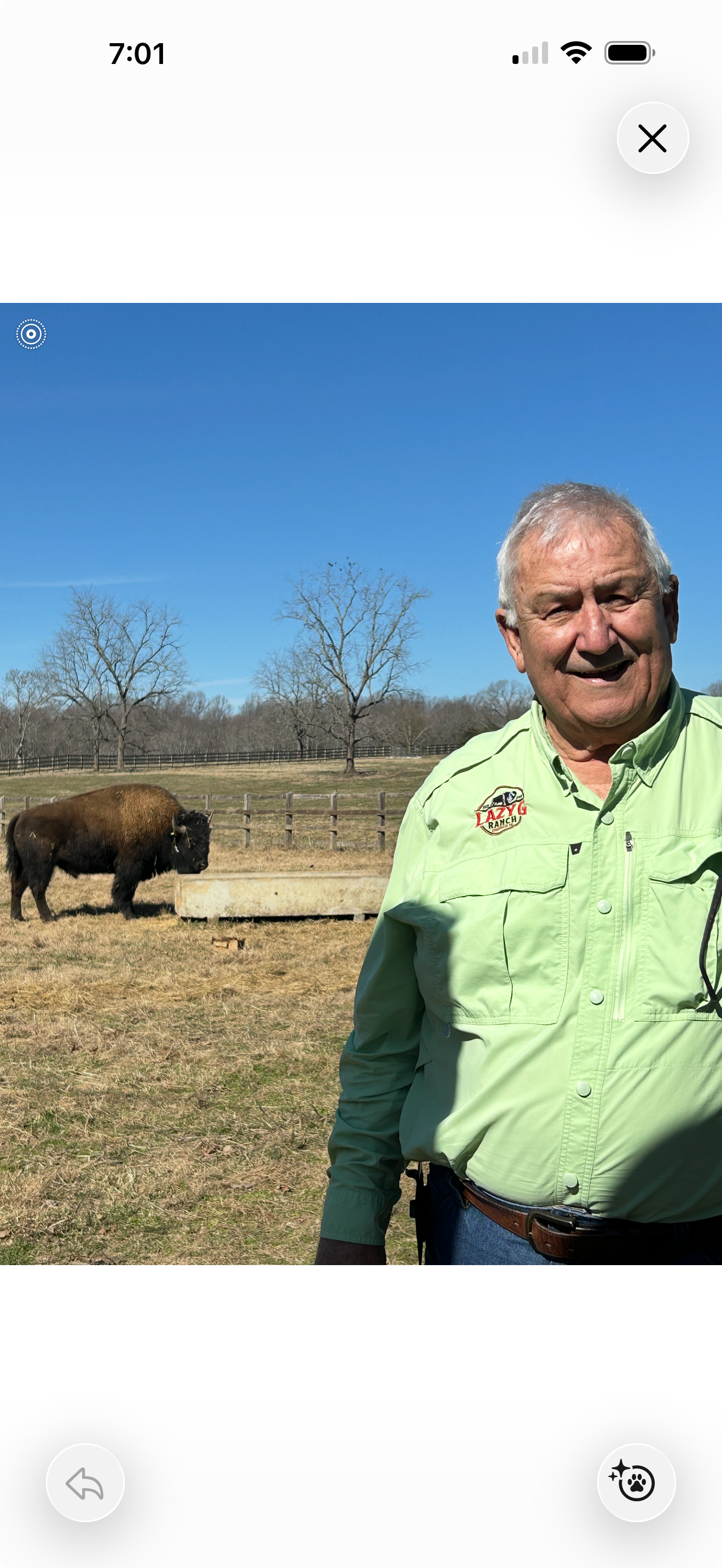 A man smiling outdoors on a farm, wearing a light green shirt with a logo that says Lazy G Ranch. In the background, there are leafless trees, a wooden fence, and a bison standing near a feeding trough under a clear blue sky.