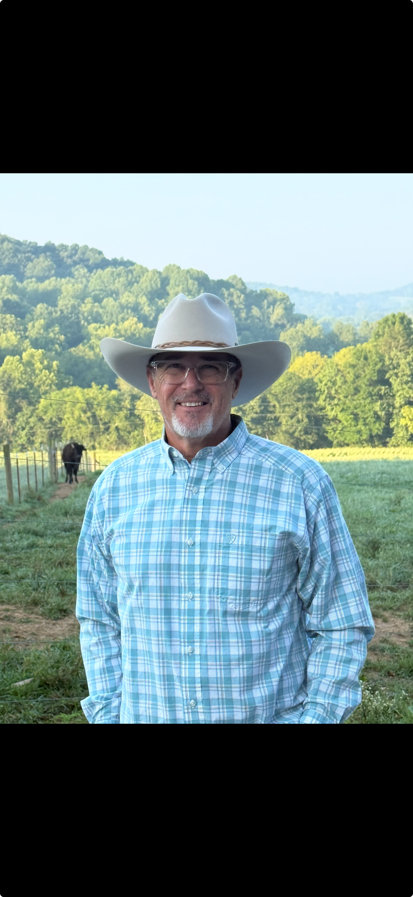 A smiling man wearing glasses, a light blue plaid shirt, and a white cowboy hat standing outdoors in a rural setting with green fields and rolling hills in the background.