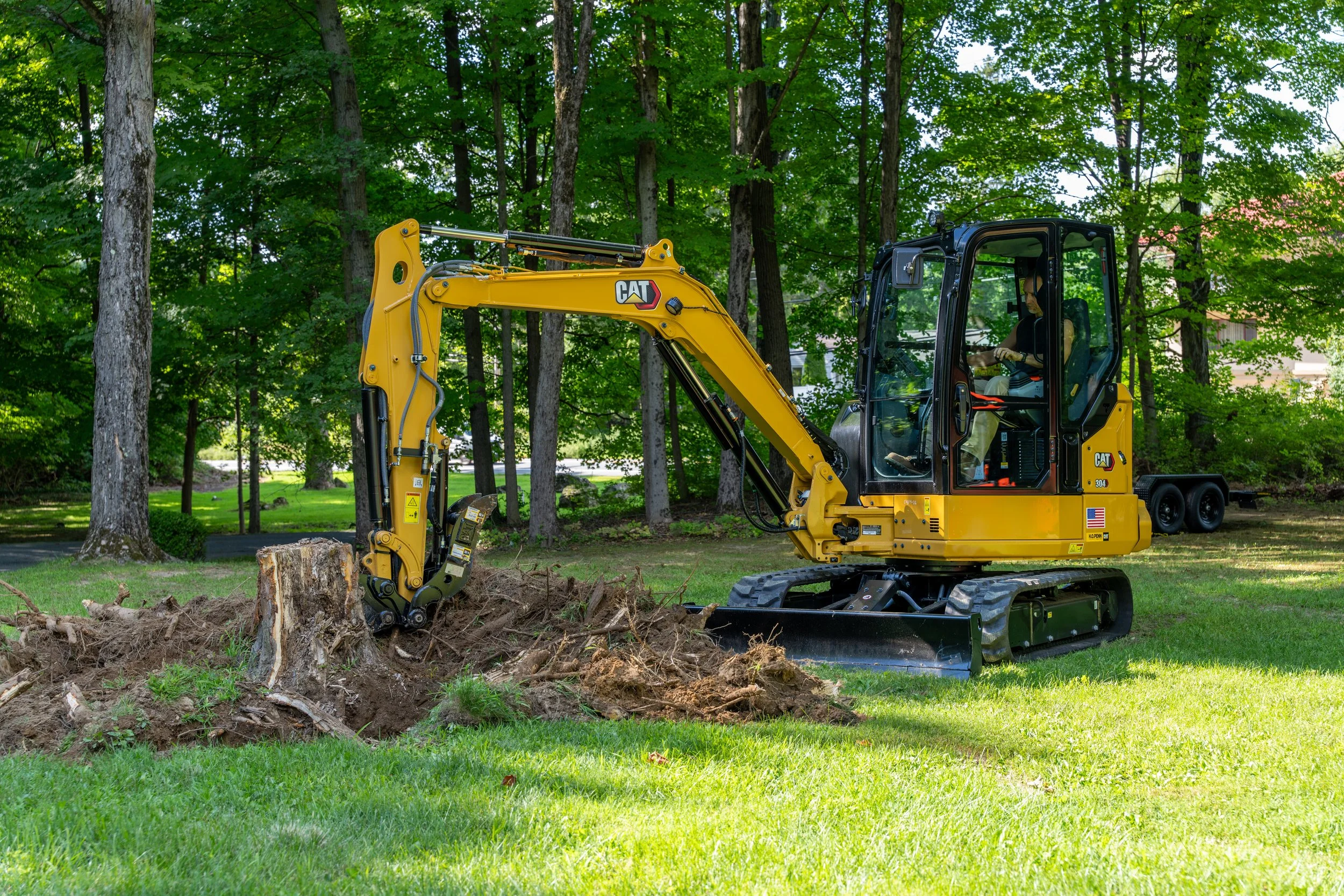 A yellow Caterpillar mini excavator digging in a grassy area surrounded by tall green trees.
