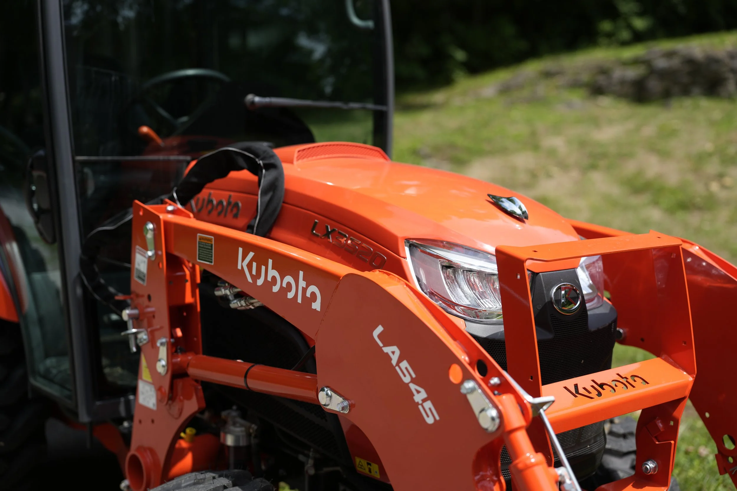 Close-up of an orange Kubota LX Series tractor with black trim, showing the front grille and headlight, parked outdoors on a grassy area with scattered rocks and trees in the background.