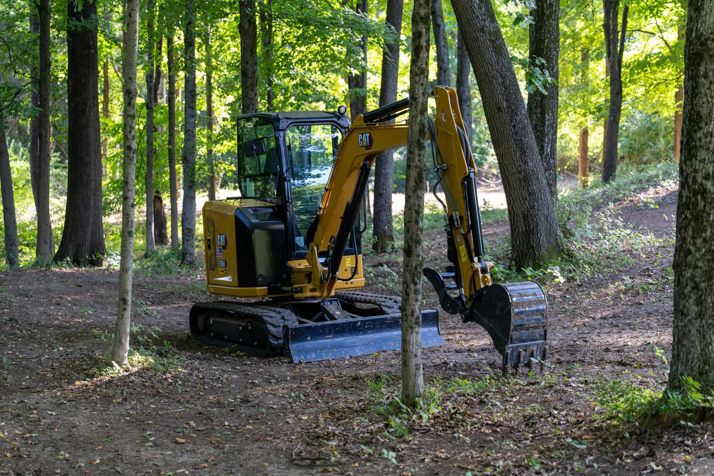 A small yellow Caterpillar excavator in a forested area with tall trees and dense green leaves in the background.