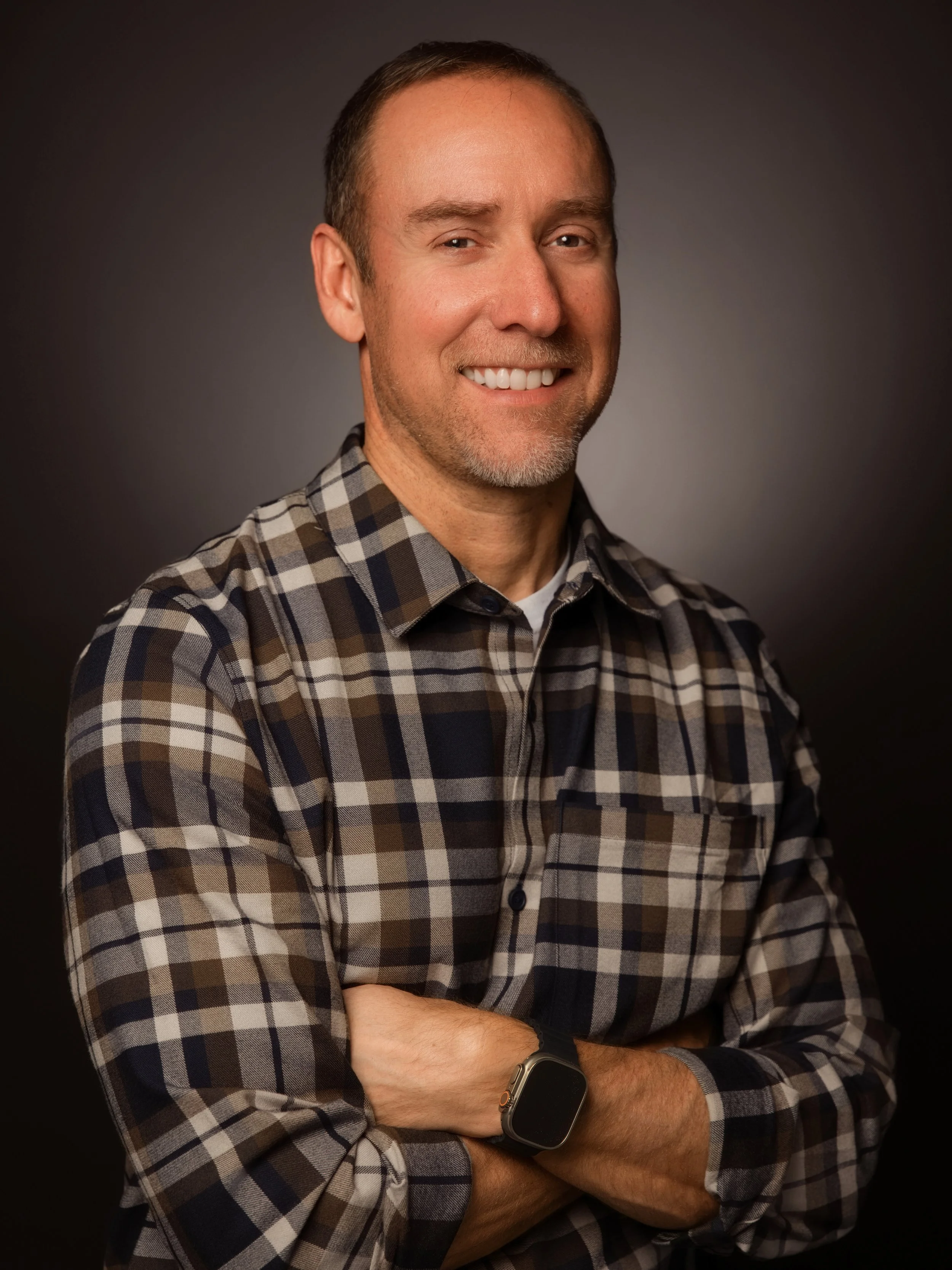 Portrait of a smiling man with short brown hair and a beard, wearing a plaid shirt and a smartwatch, standing against a dark background.