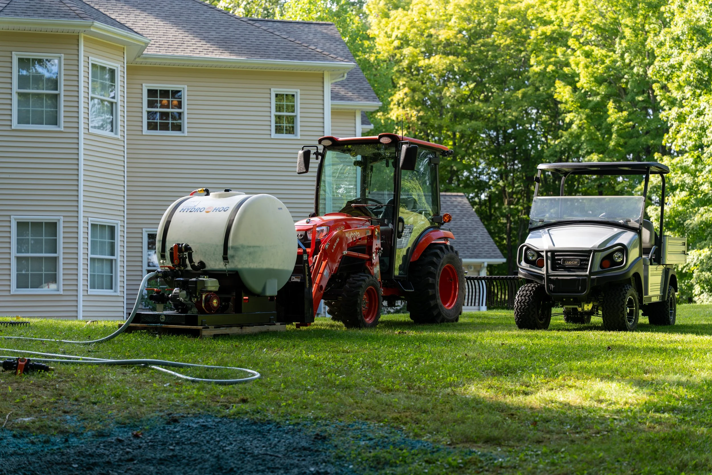 A red tractor with a large white tank attached to it, parked on a grassy yard next to a white golf cart, in front of a beige house with multiple windows and a gray roof, surrounded by green trees.