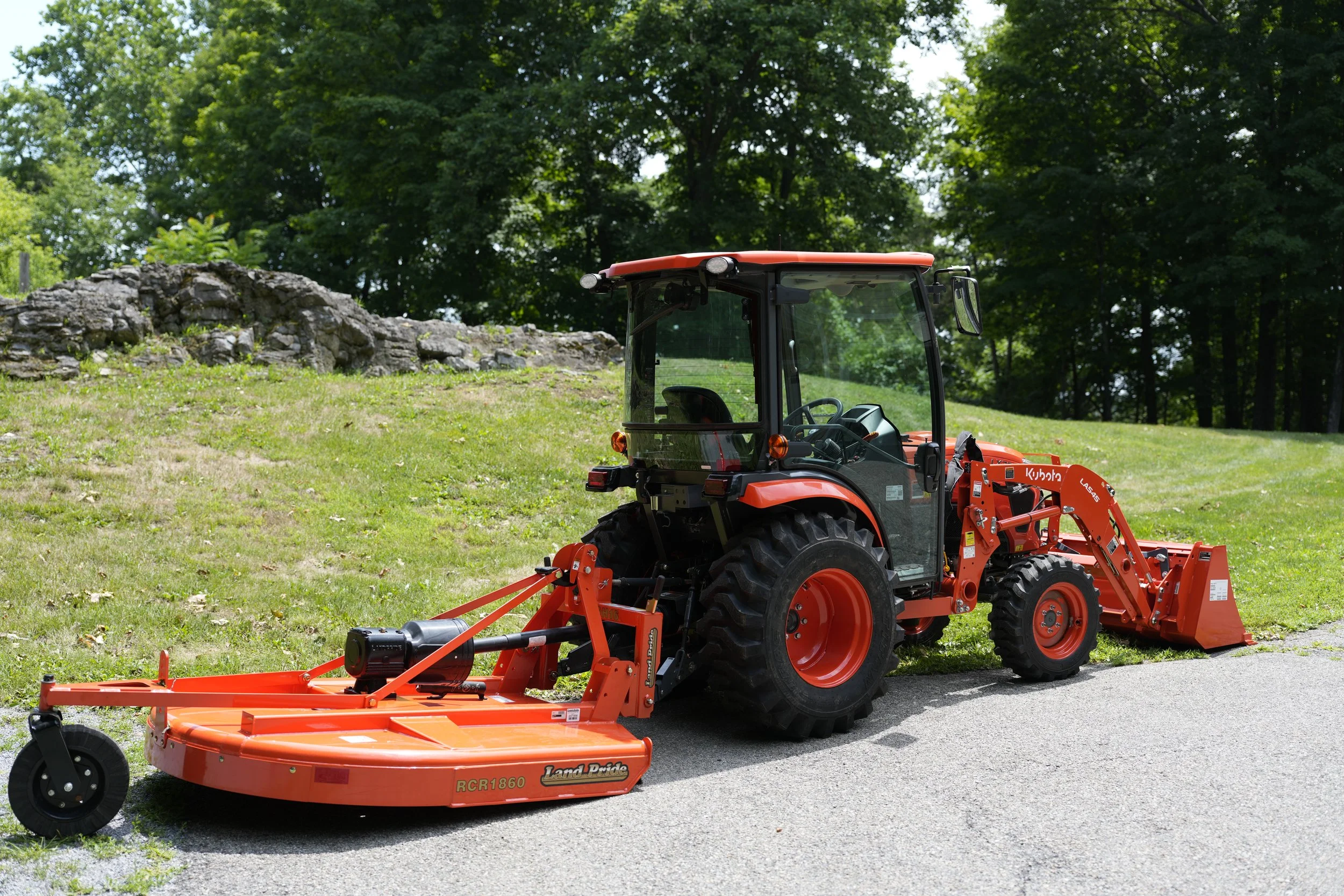Orange Kubota tractor with a large mower attached, parked on a paved road next to grassy area with stone wall and trees in the background.