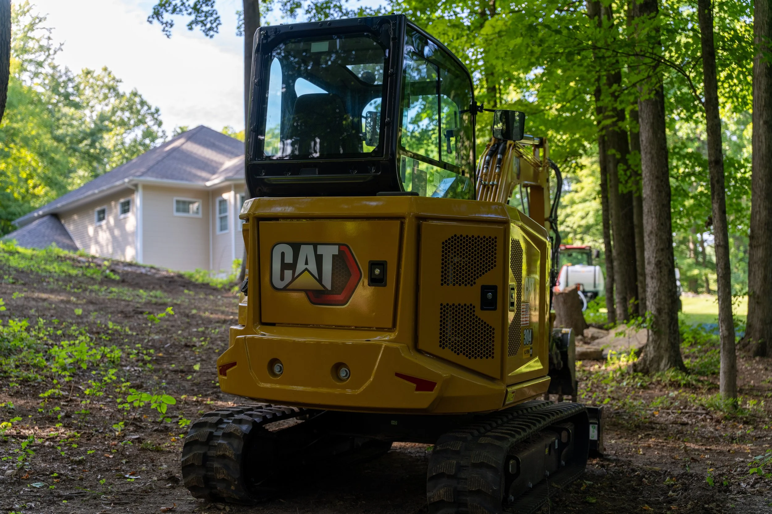 A yellow Caterpillar mini excavator on a cleared yard with trees and a house in the background.