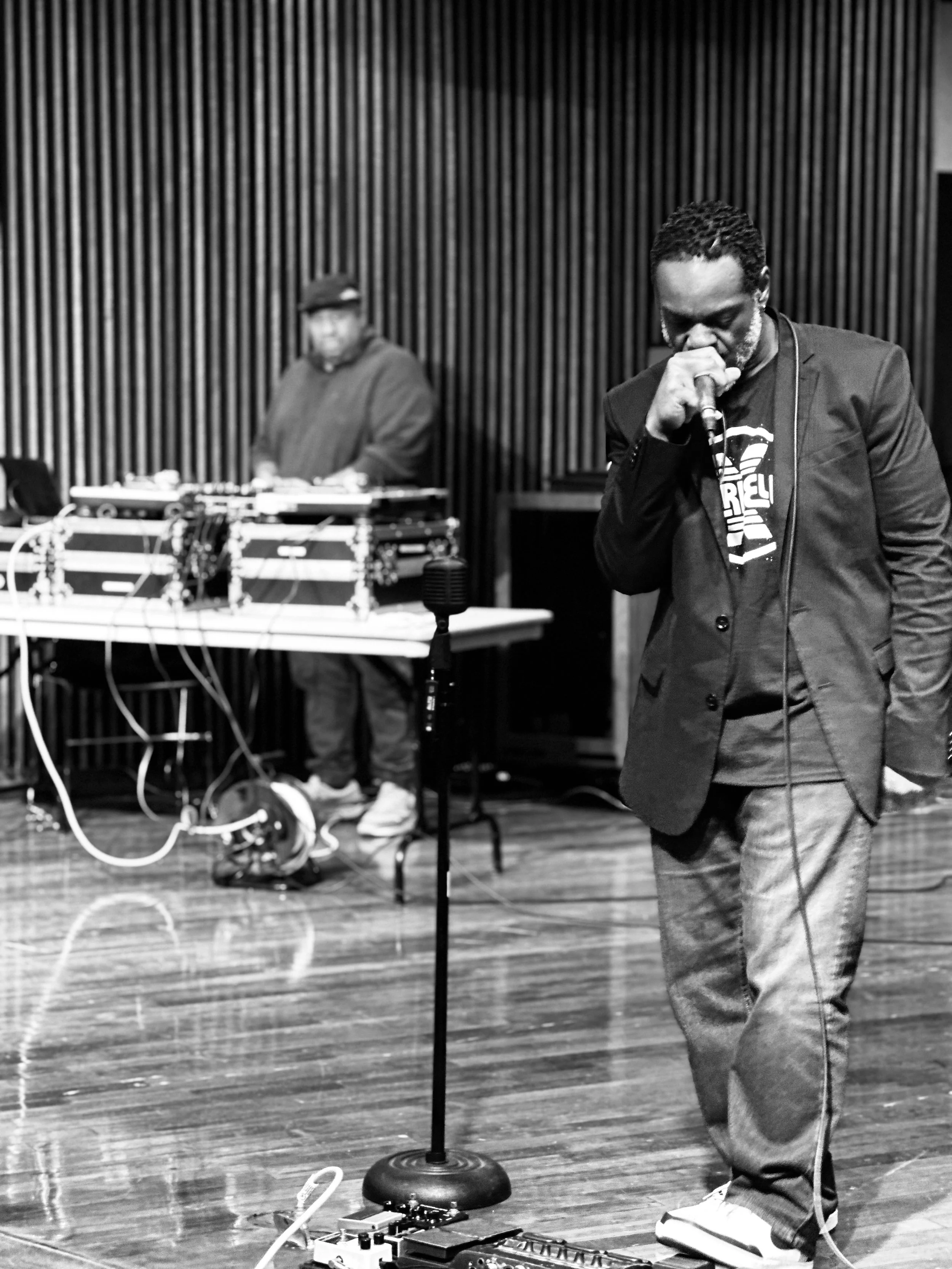 A man with a beard and dreadlocks standing in front of a microphone, with a DJ in the background operating turntables and equipment, on a wooden stage with a vertical wood-paneled wall behind them.