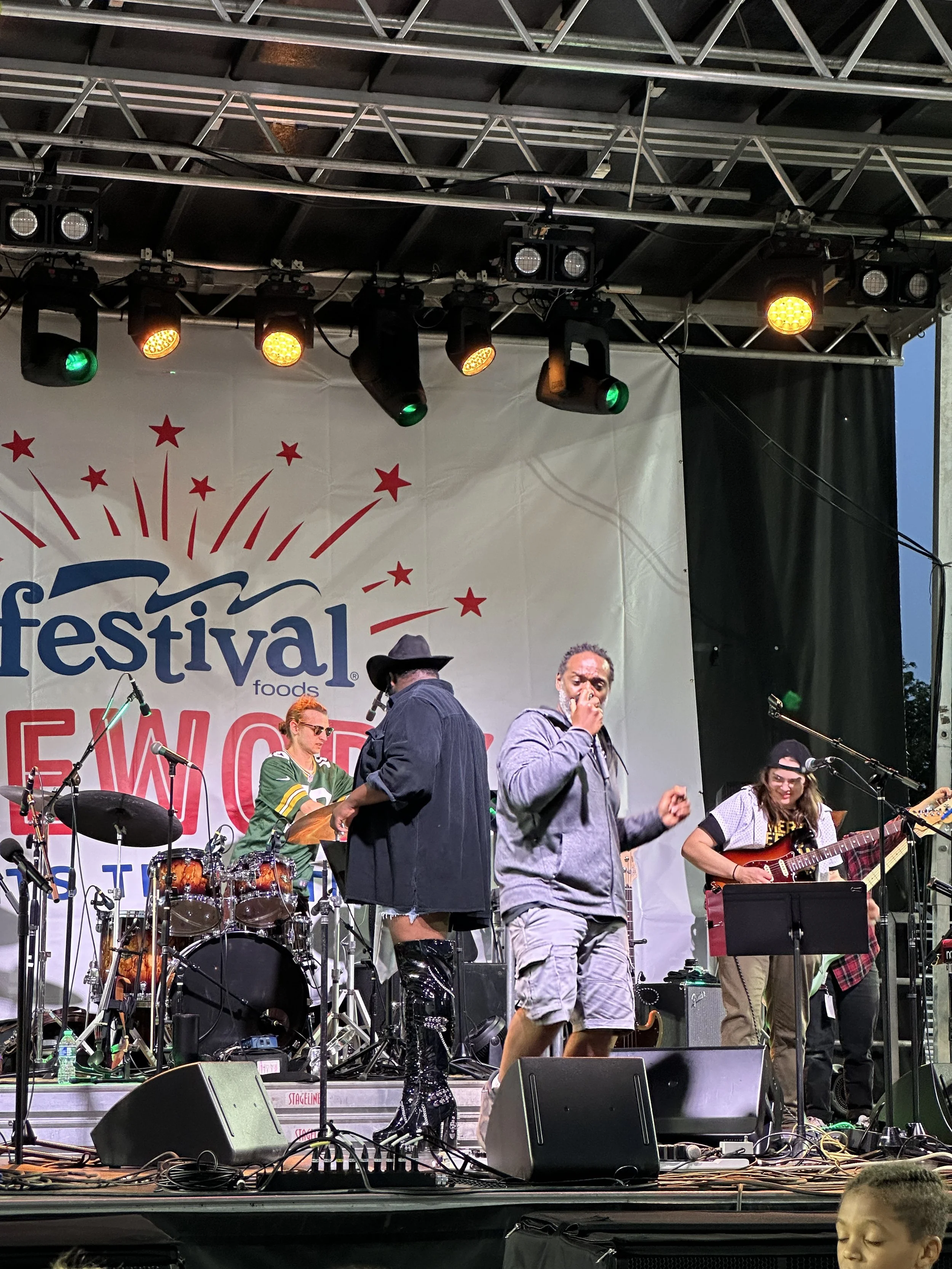 People performing on an outdoor stage at a festival, with musical instruments and sound equipment visible, under stage lights.