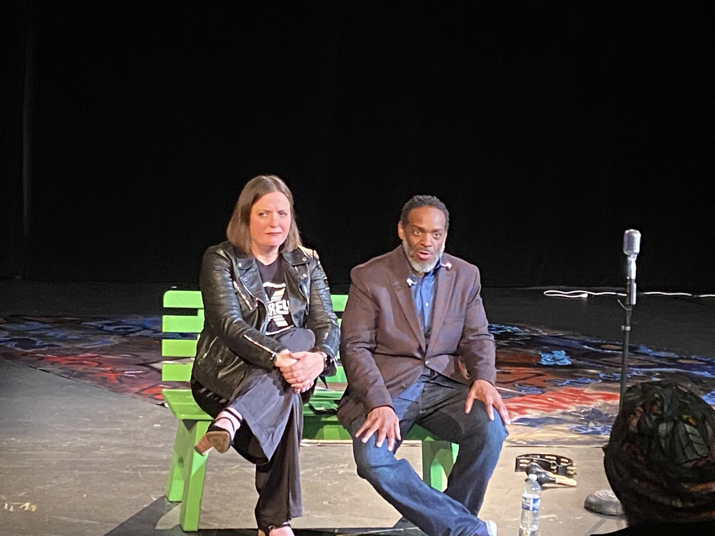 Two individuals seated on a stage with a microphone and water bottles, against a black background. One person is a woman with shoulder-length hair, wearing a leather jacket, and the other is a man with short hair and a beard, wearing a blazer.