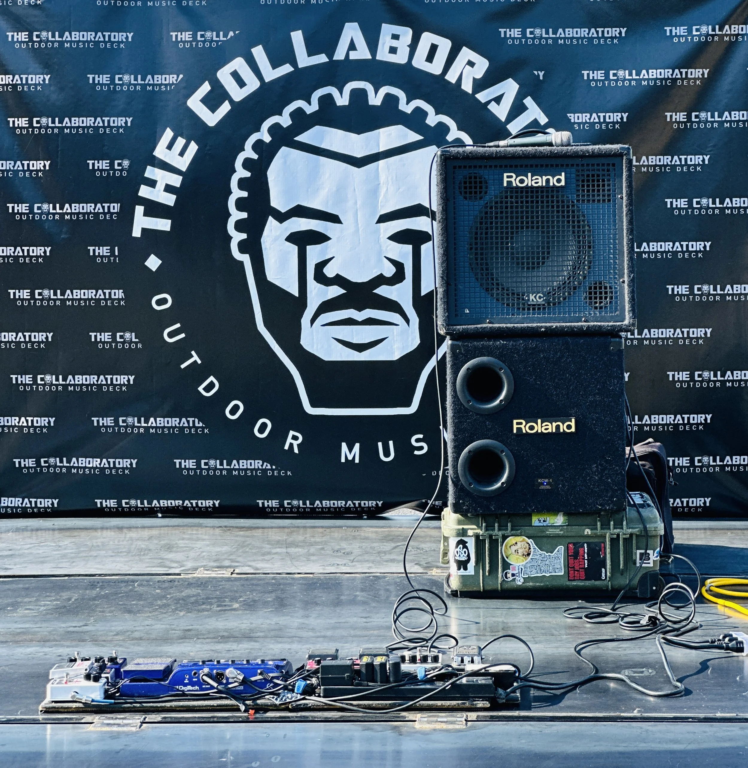 A music stage setup featuring a blue speaker, pedalboard, and power cords in front of a backdrop with the logo and text indicating The Laboratory outdoor music deck.